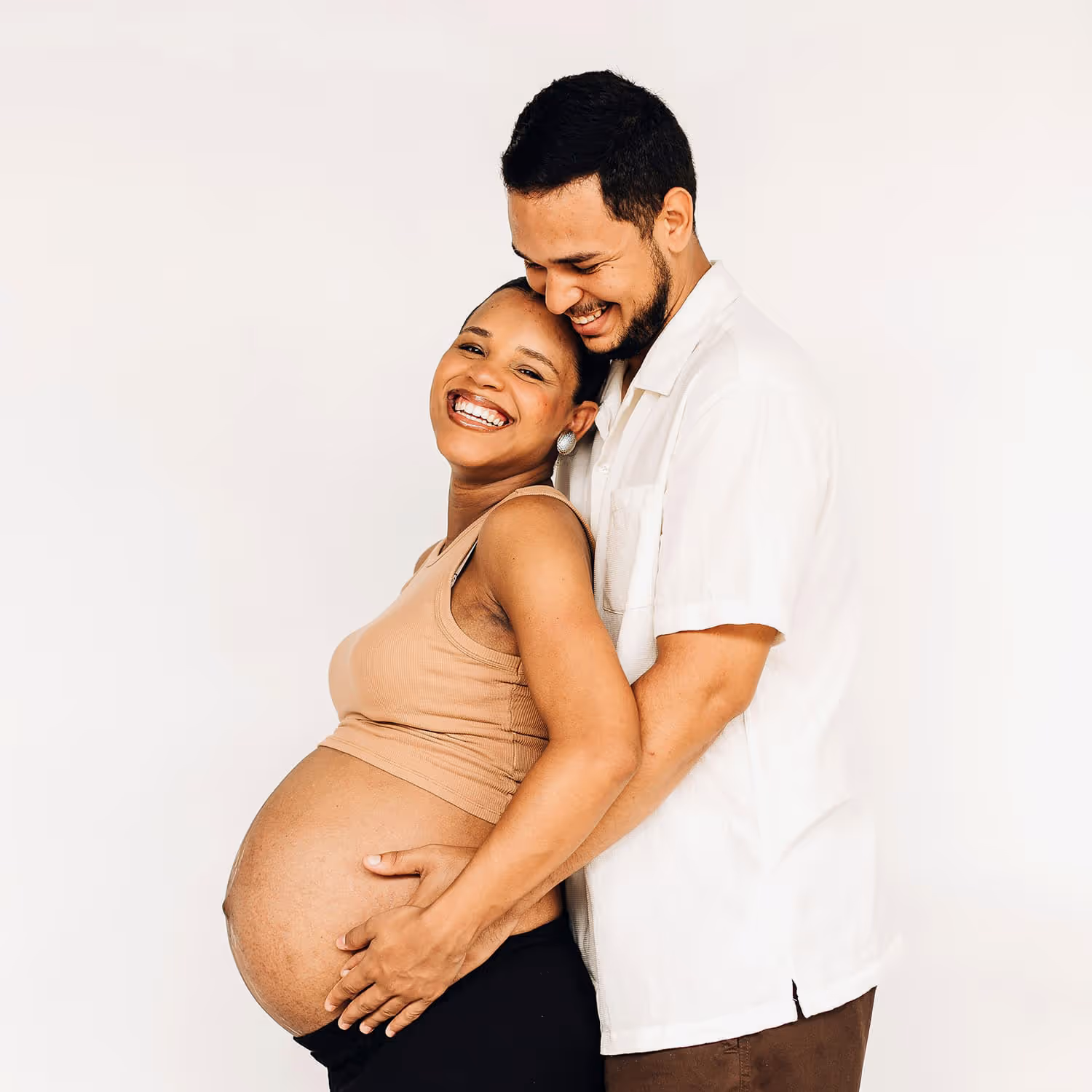 Smiling pregnant person and partner embracing against a neutral background, both with hands gently resting on the pregnant belly