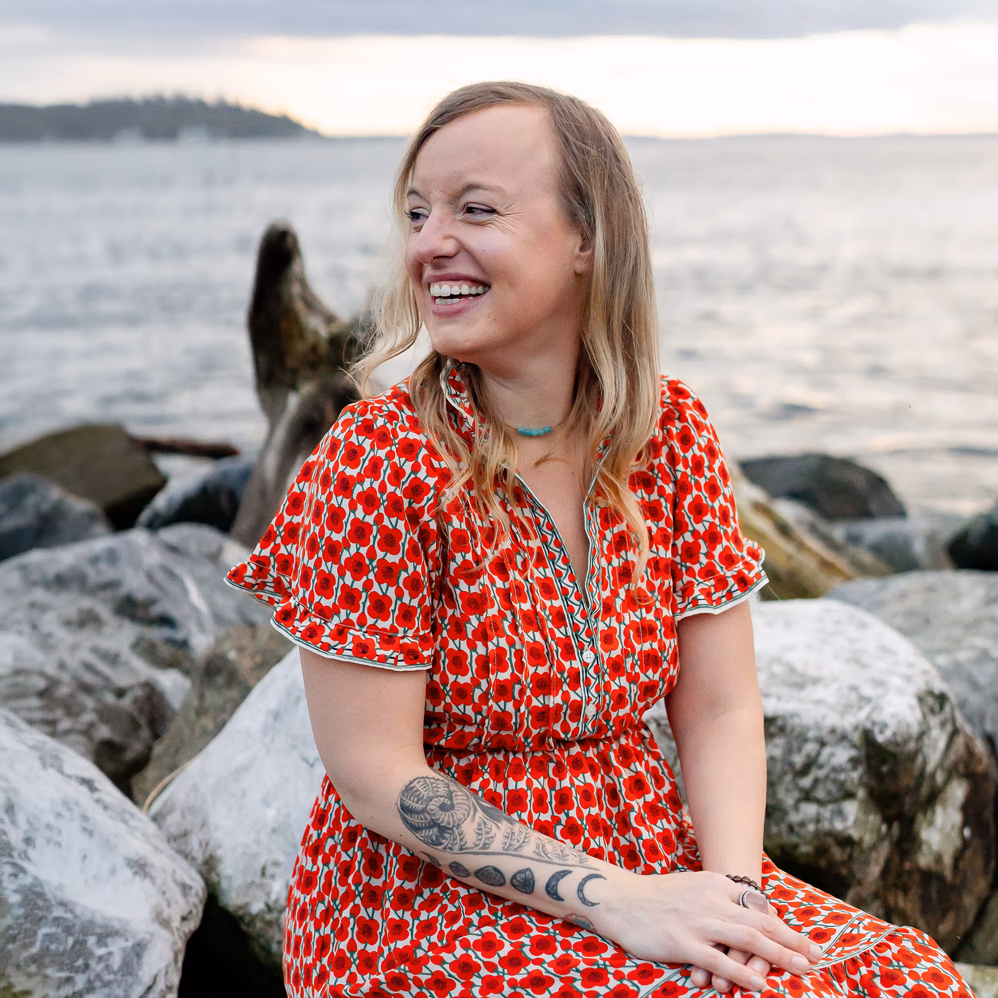 Woman sitting on driftwood on a rocky beach, looking over her right shoulder and smiling.