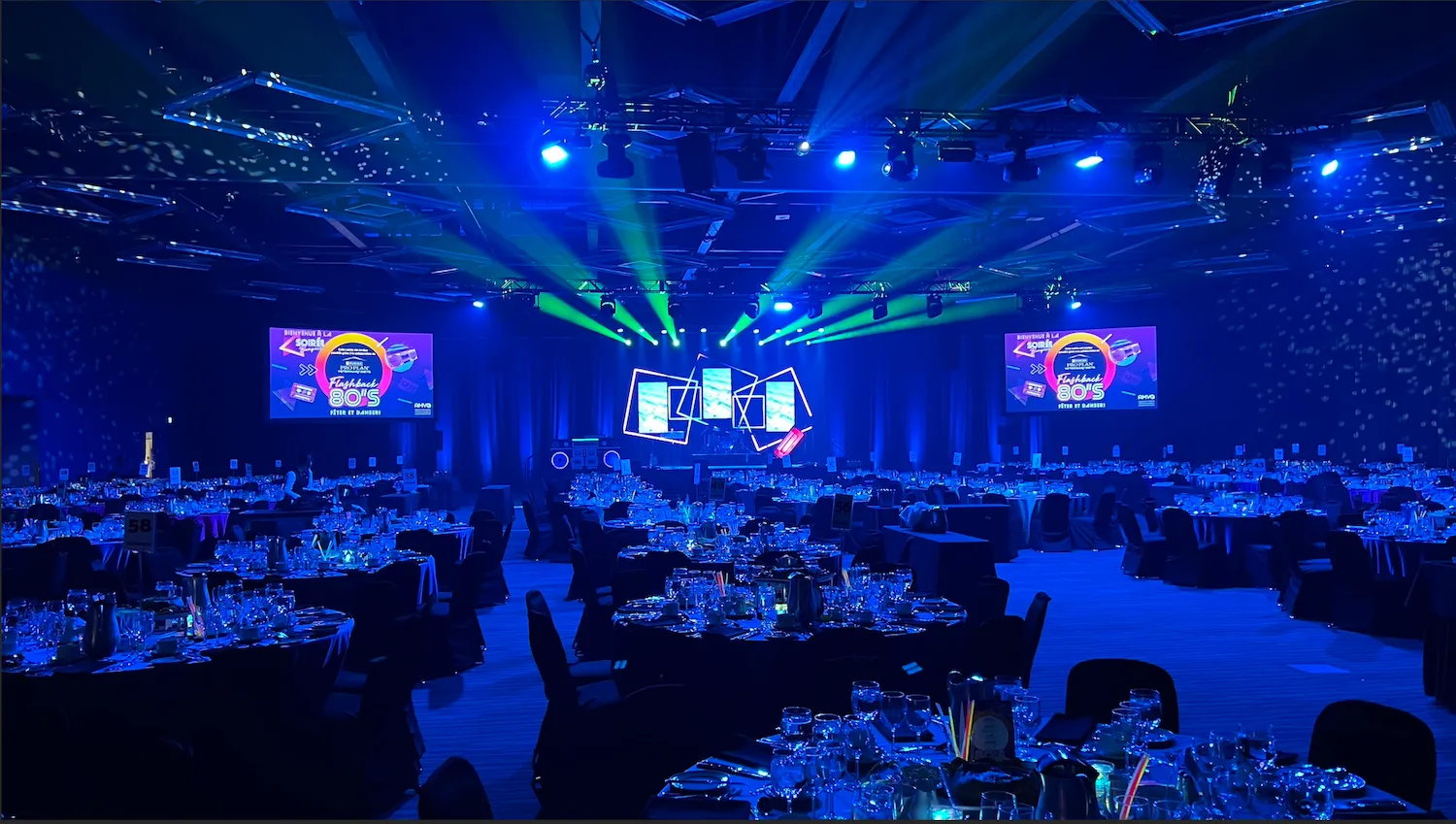 Grande salle de banquet illuminée en bleu avec écrans et tables dressées.