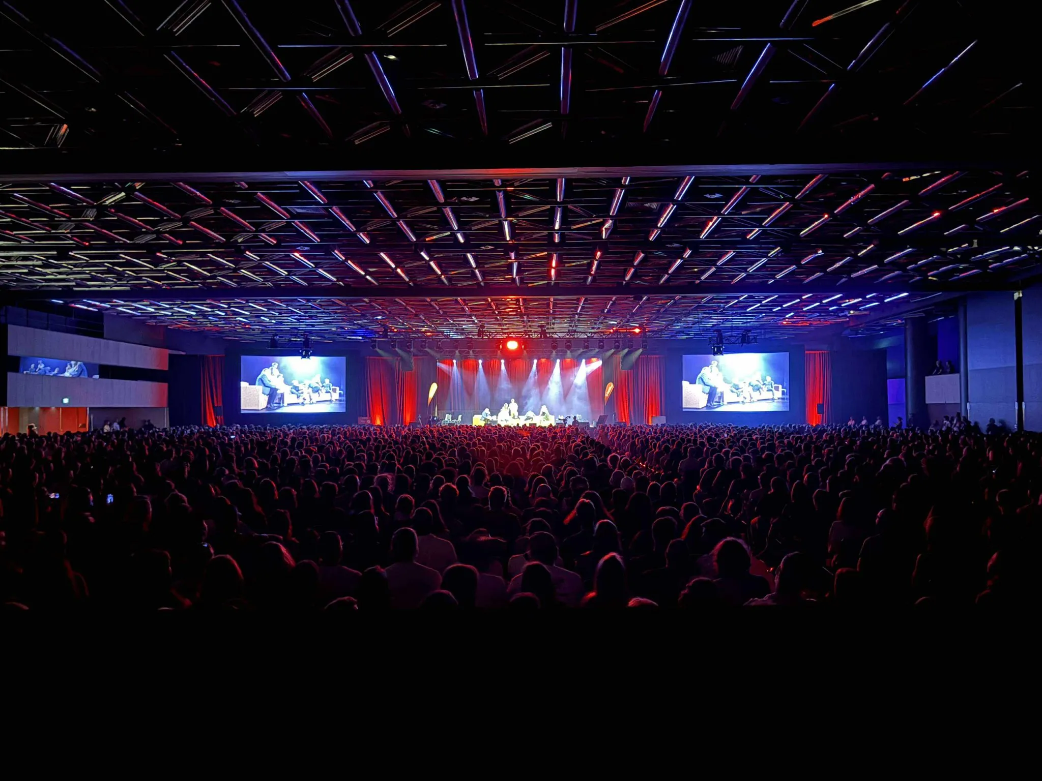Grande salle de concert avec foule illuminée par des lumières bleues et rouges au plafond.