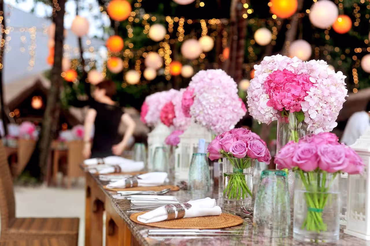 Elegant outdoor catering setup with pink and purple flower centerpieces, decorative lighting, and rustic charm for a festive event.