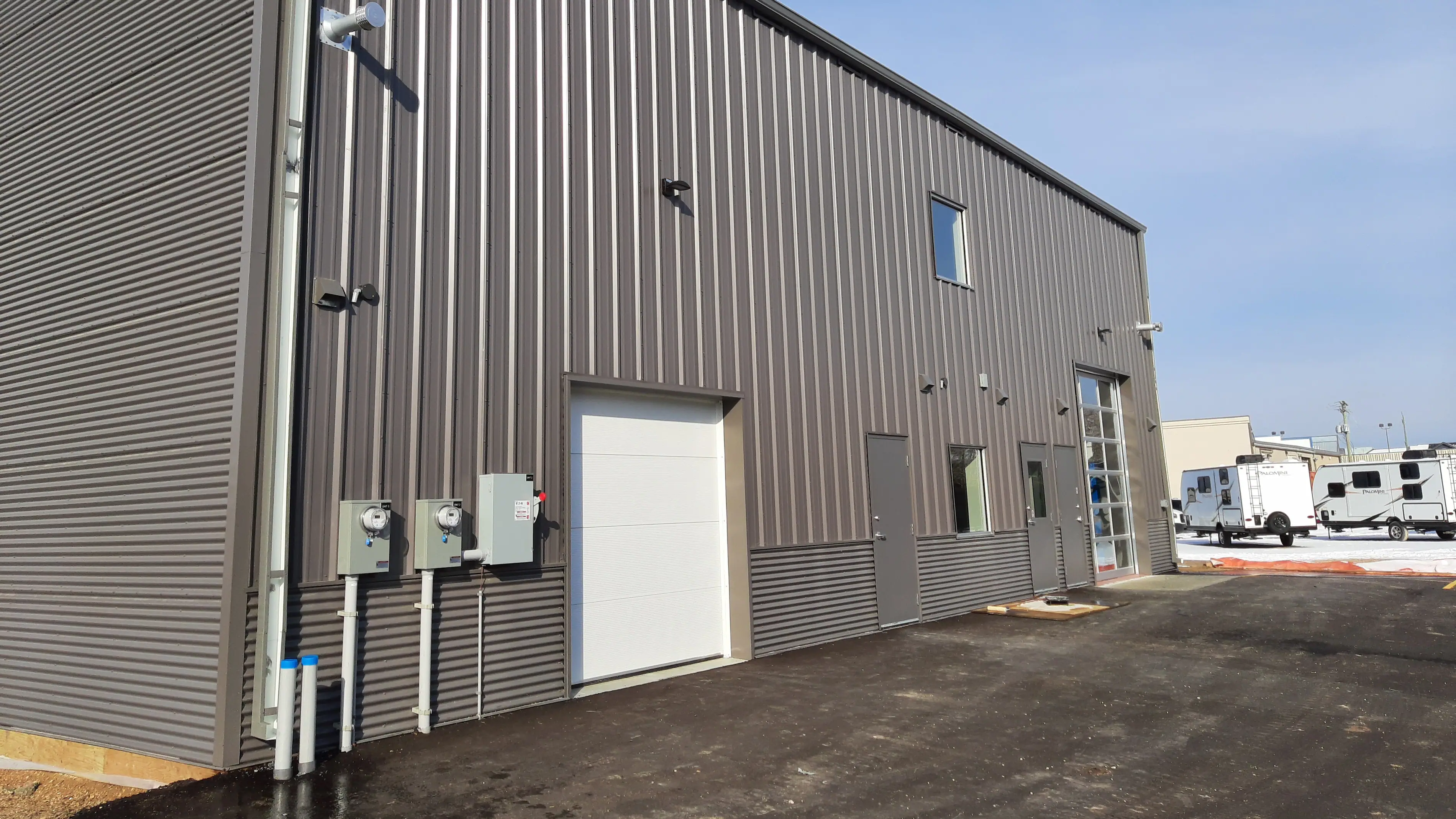 A modern industrial building with gray corrugated metal siding, a white garage door, and several windows. Bright, clear day with RVs in the background.