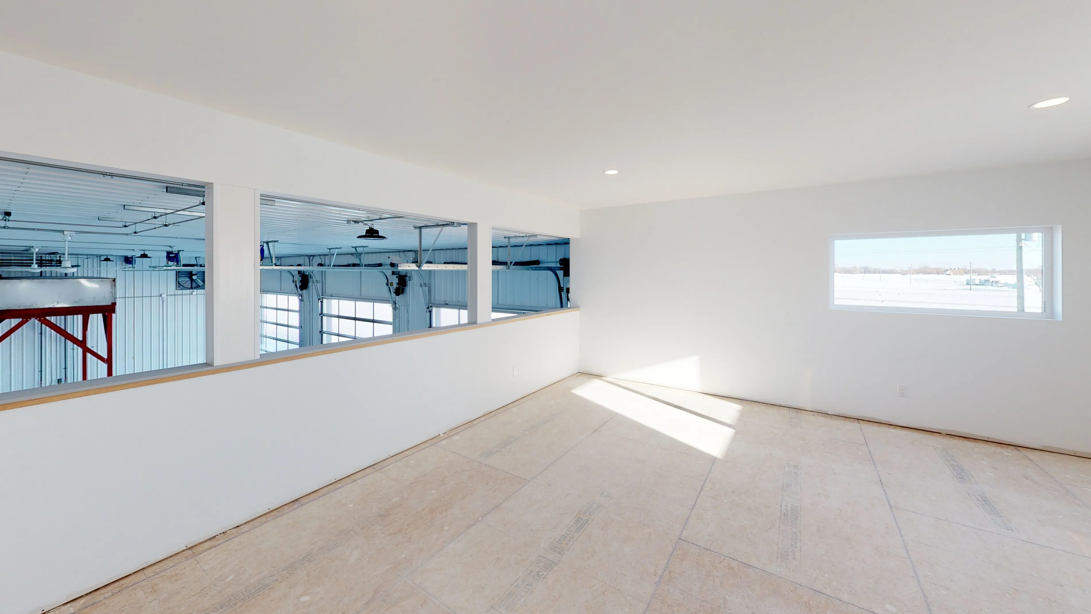 A spacious, sunlit room with light wooden floors and white walls. A window and interior glass panels overlook an industrial space, creating a clean, open feel.