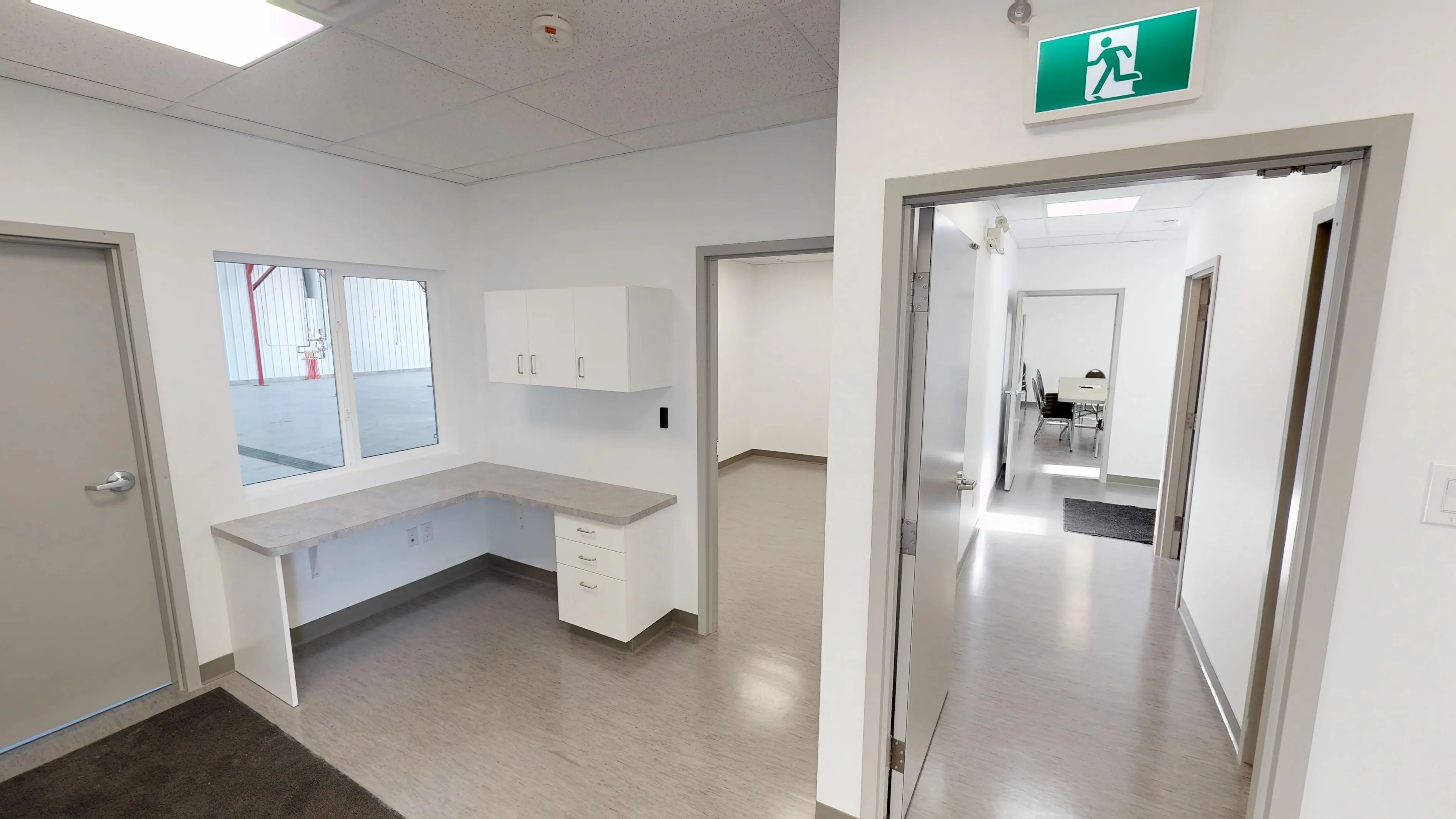 A clean, modern office with white walls and gray flooring. It features a window, a desk with cabinets, and a green exit sign above a hallway entrance.