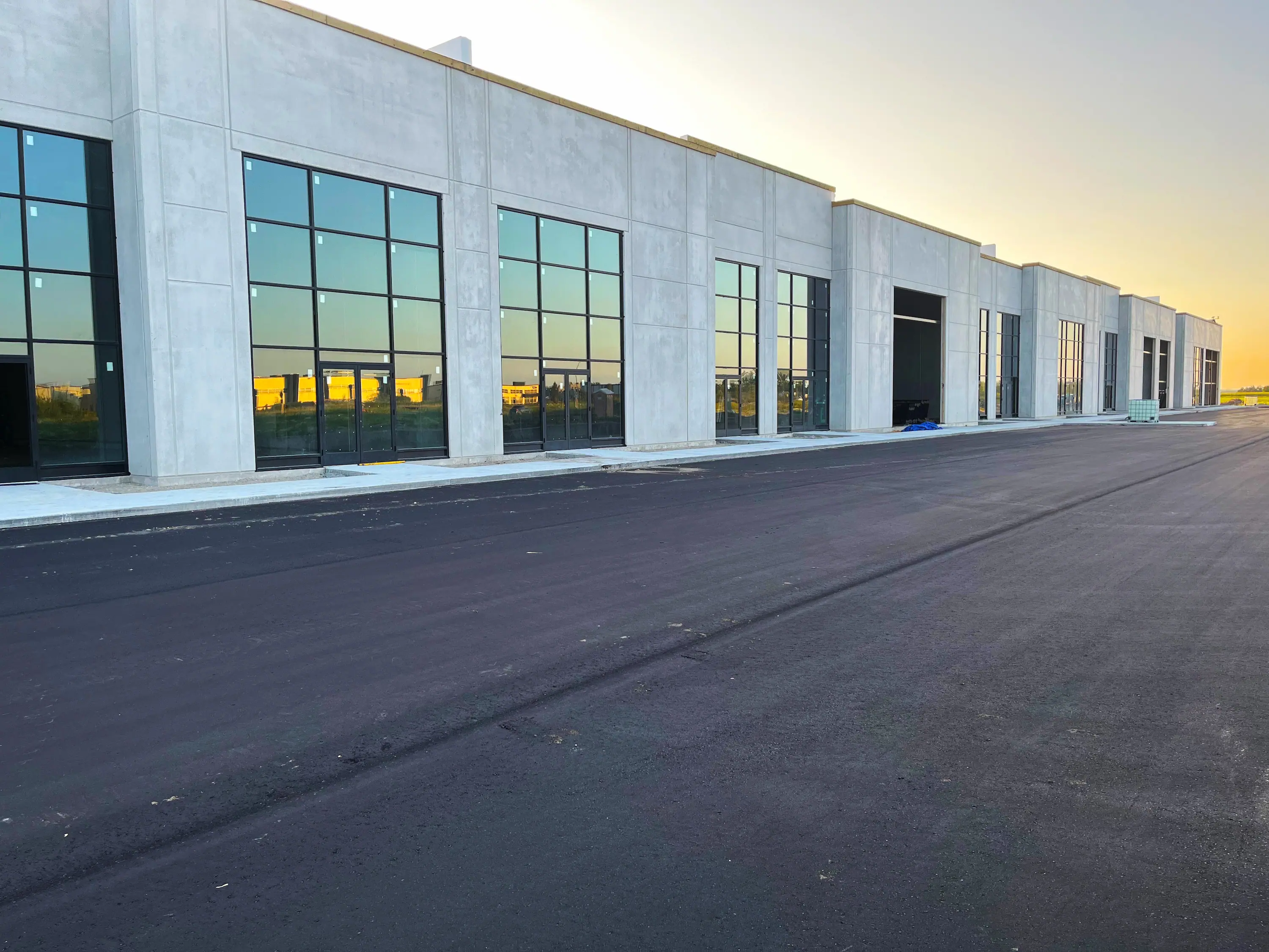 Modern incomplete industrial building with large reflective windows alongside an empty paved road. The sky is clear, creating a tranquil sunset ambiance.