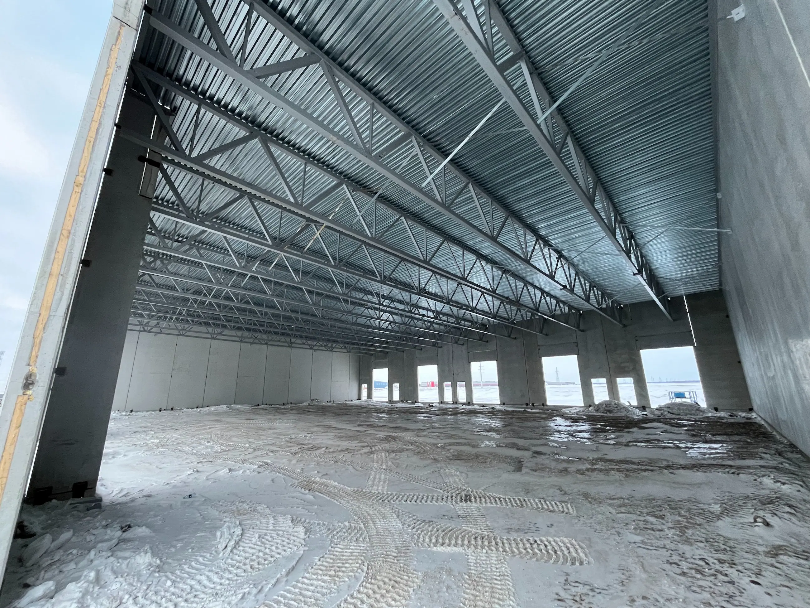Interior view of an unfinished warehouse with exposed steel beams and a partially snow-covered floor. Bright, cold atmosphere with large window openings.