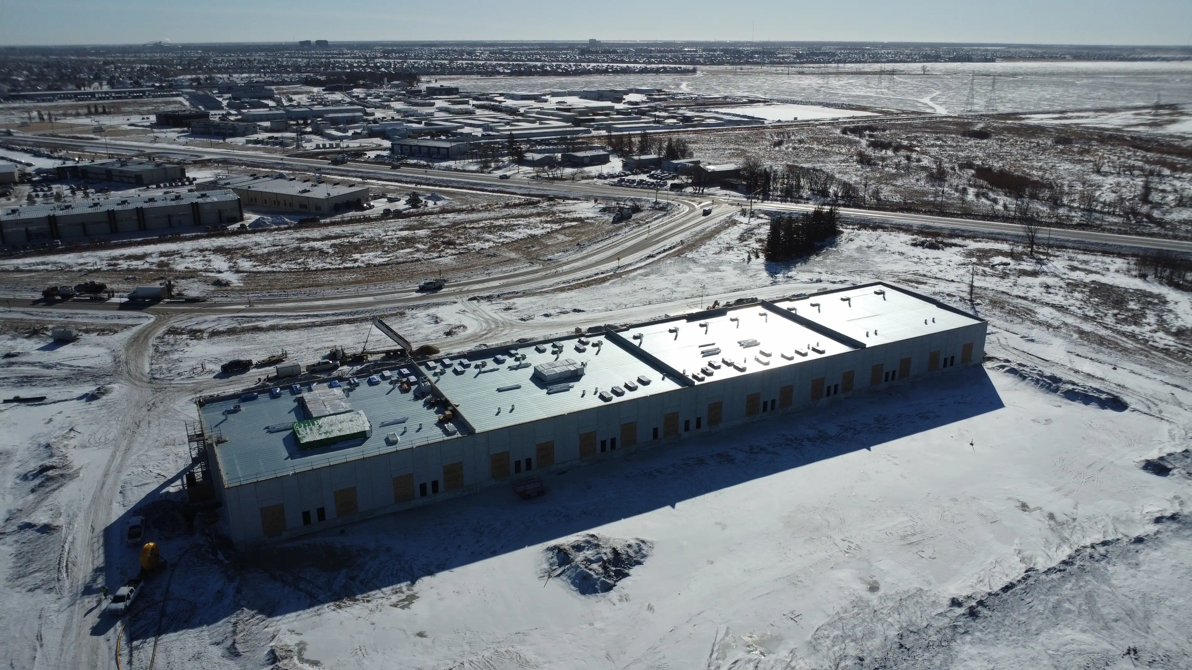 Aerial view of a large industrial building with a reflective roof in a snowy landscape. Surrounding the structure are snow-covered fields and distant cityscape.