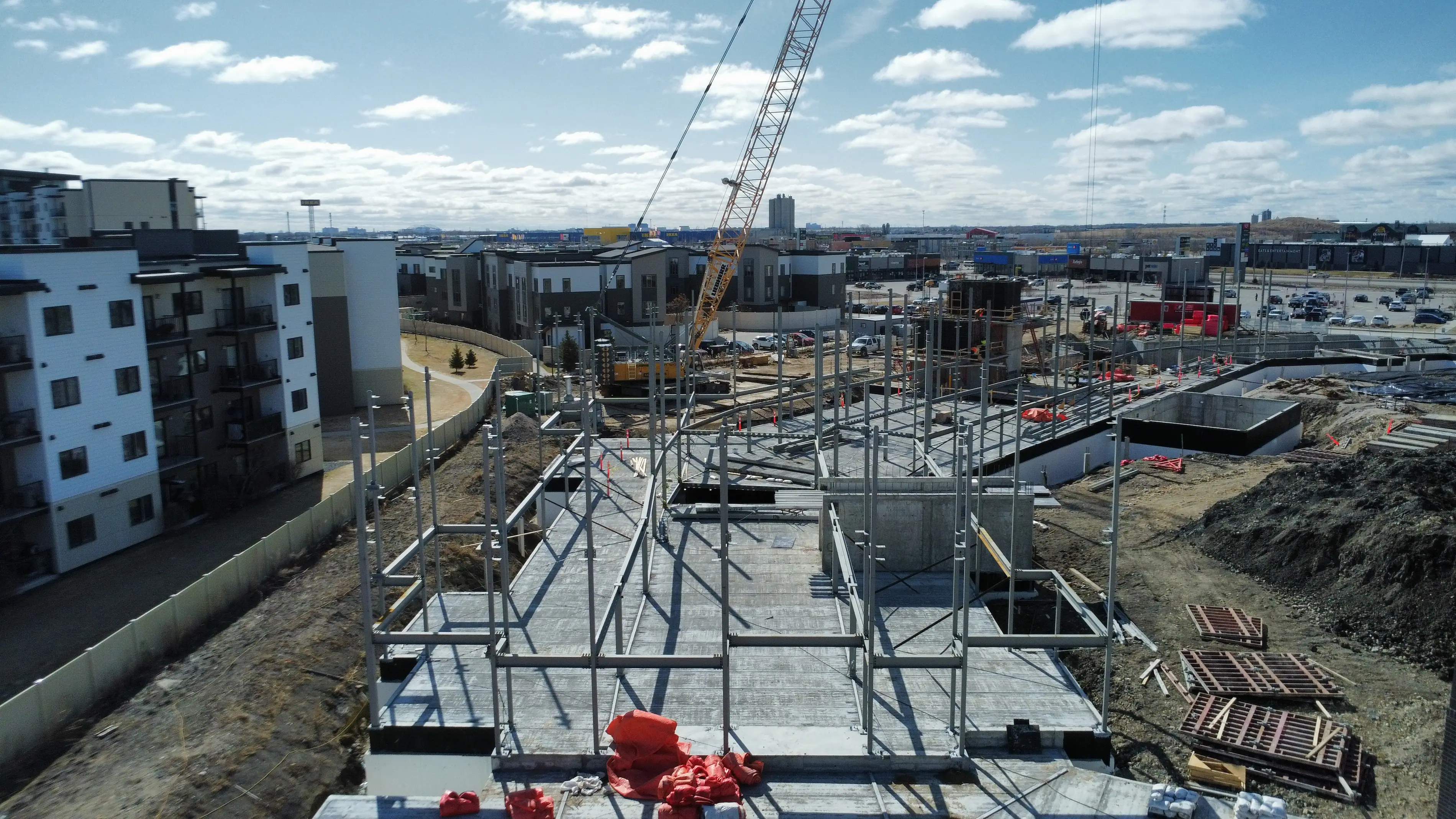 Steel framework and concrete foundation of a building under construction, surrounded by equipment, materials, and nearby residential buildings on a partly cloudy day.