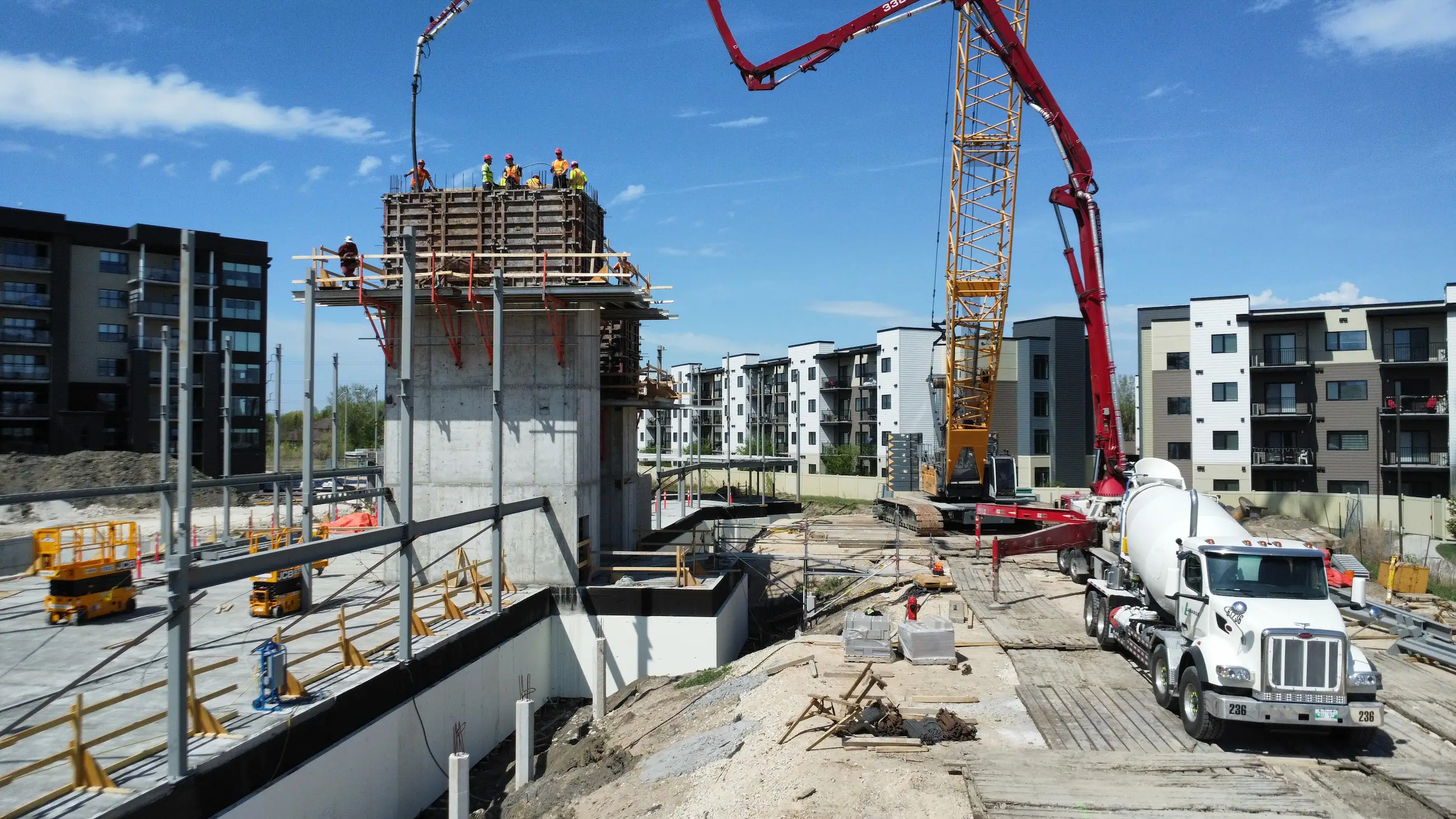 Construction workers stand atop a concrete structure while a concrete pump truck pours material; nearby, a crane and other construction equipment are visible on site.