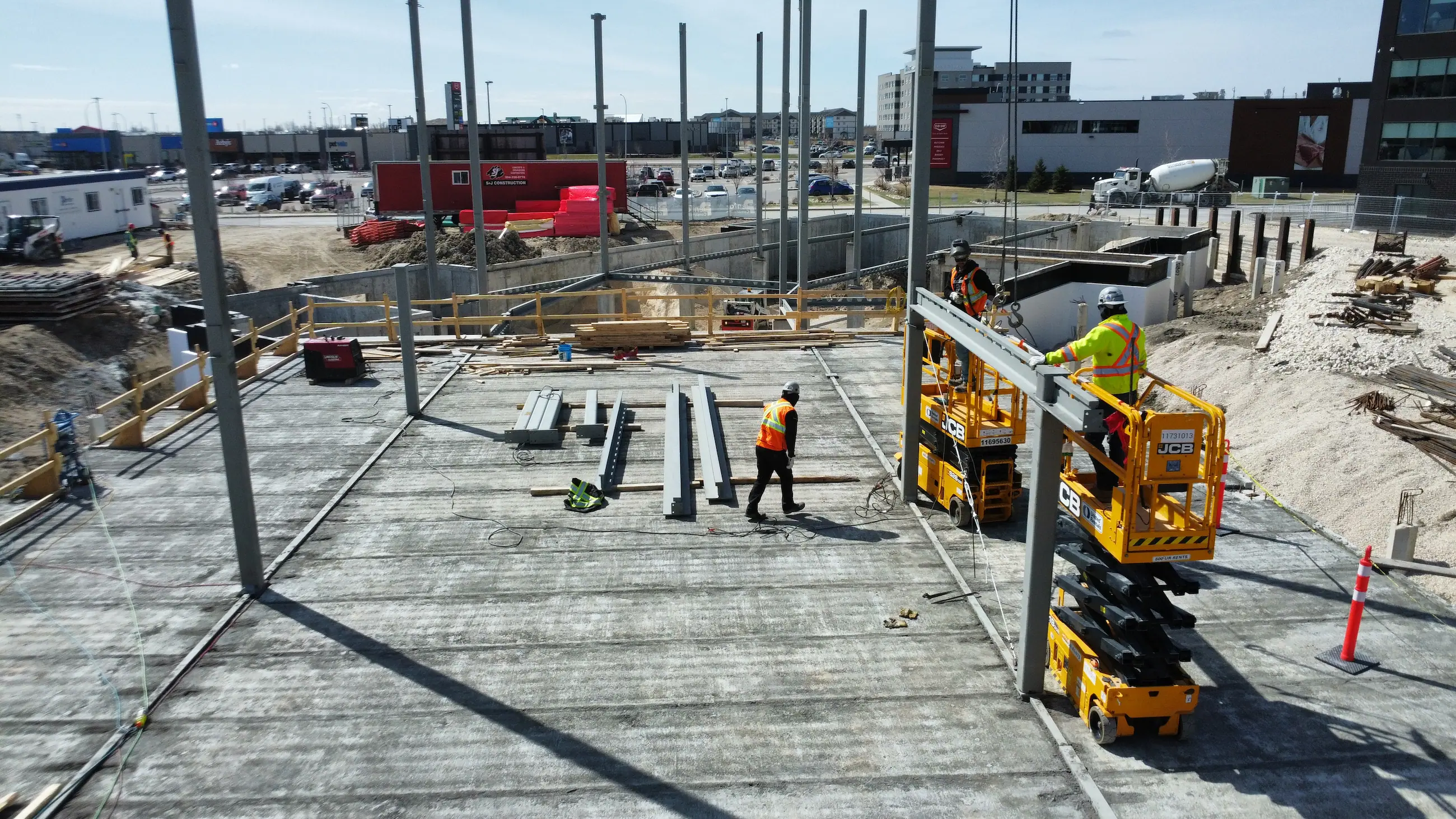 A construction site with workers in safety gear on a concrete foundation, using yellow machinery to position steel beams. Urban setting with buildings visible.