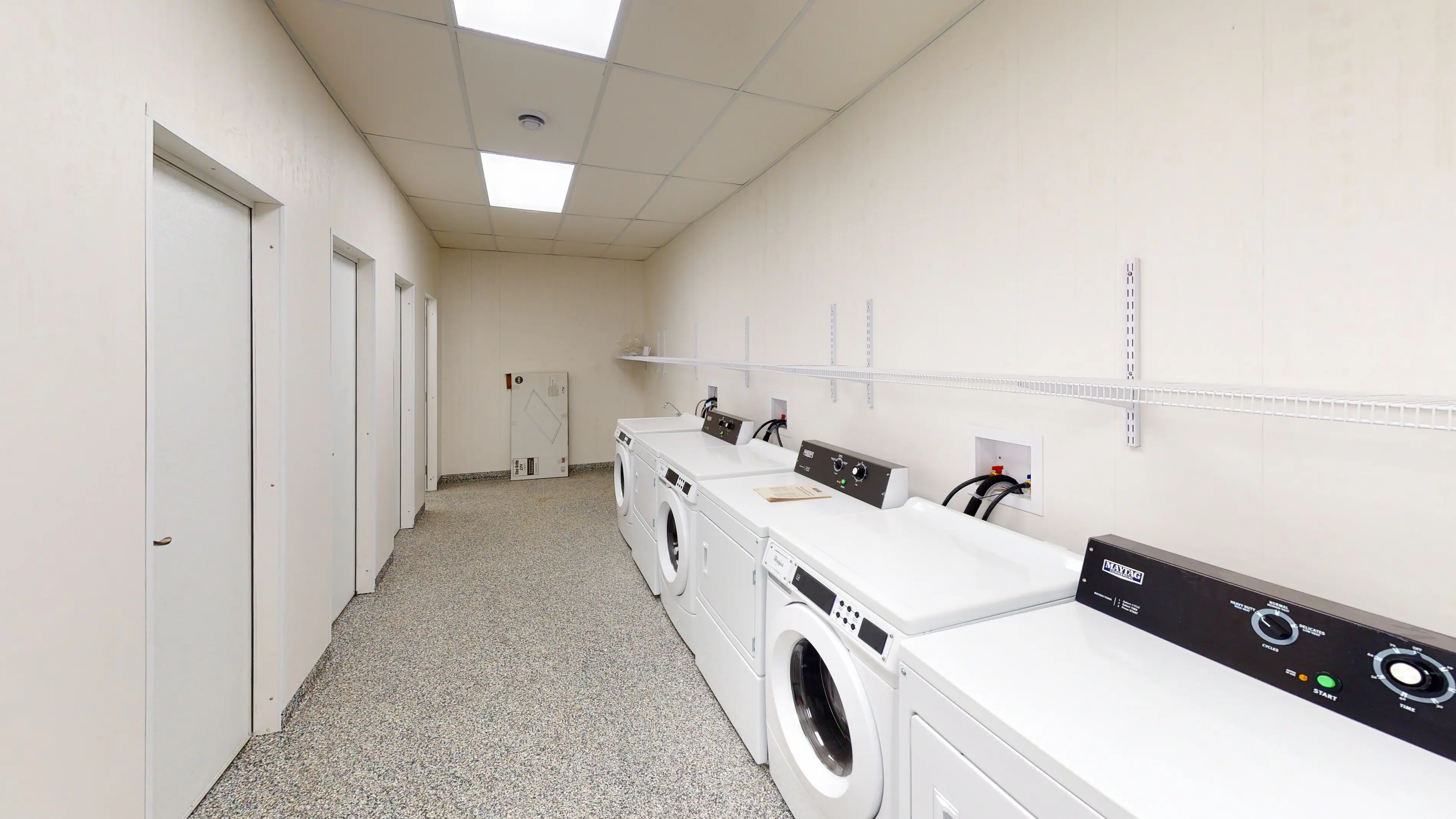 Brightly lit laundry room with a row of white washers and dryers on the right. Beige walls, speckled floor, and three closed doors on the left. Clean and orderly.