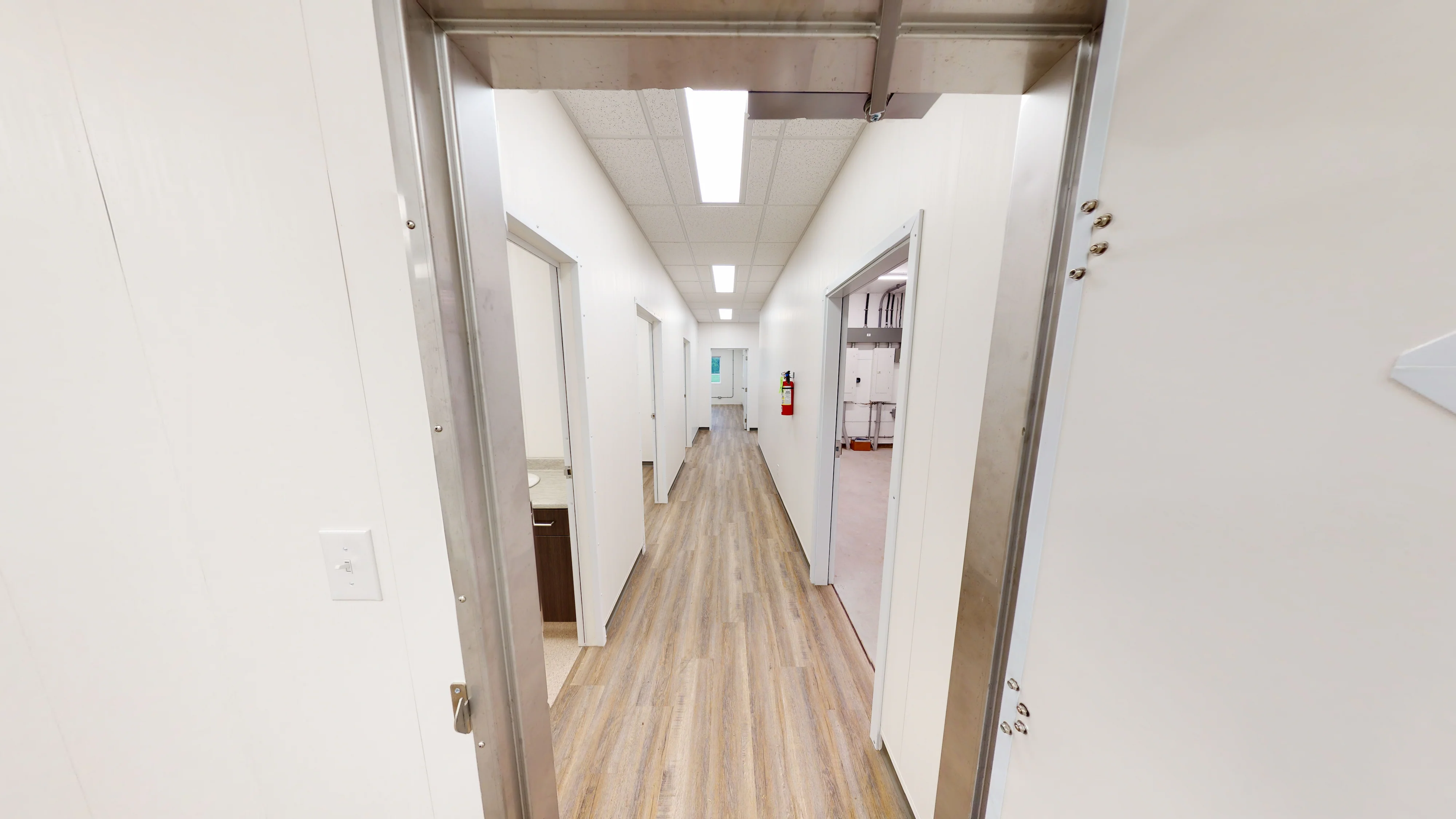 A long corridor with white walls, wooden flooring, and multiple doors on each side, leading to a well-lit room at the far end.