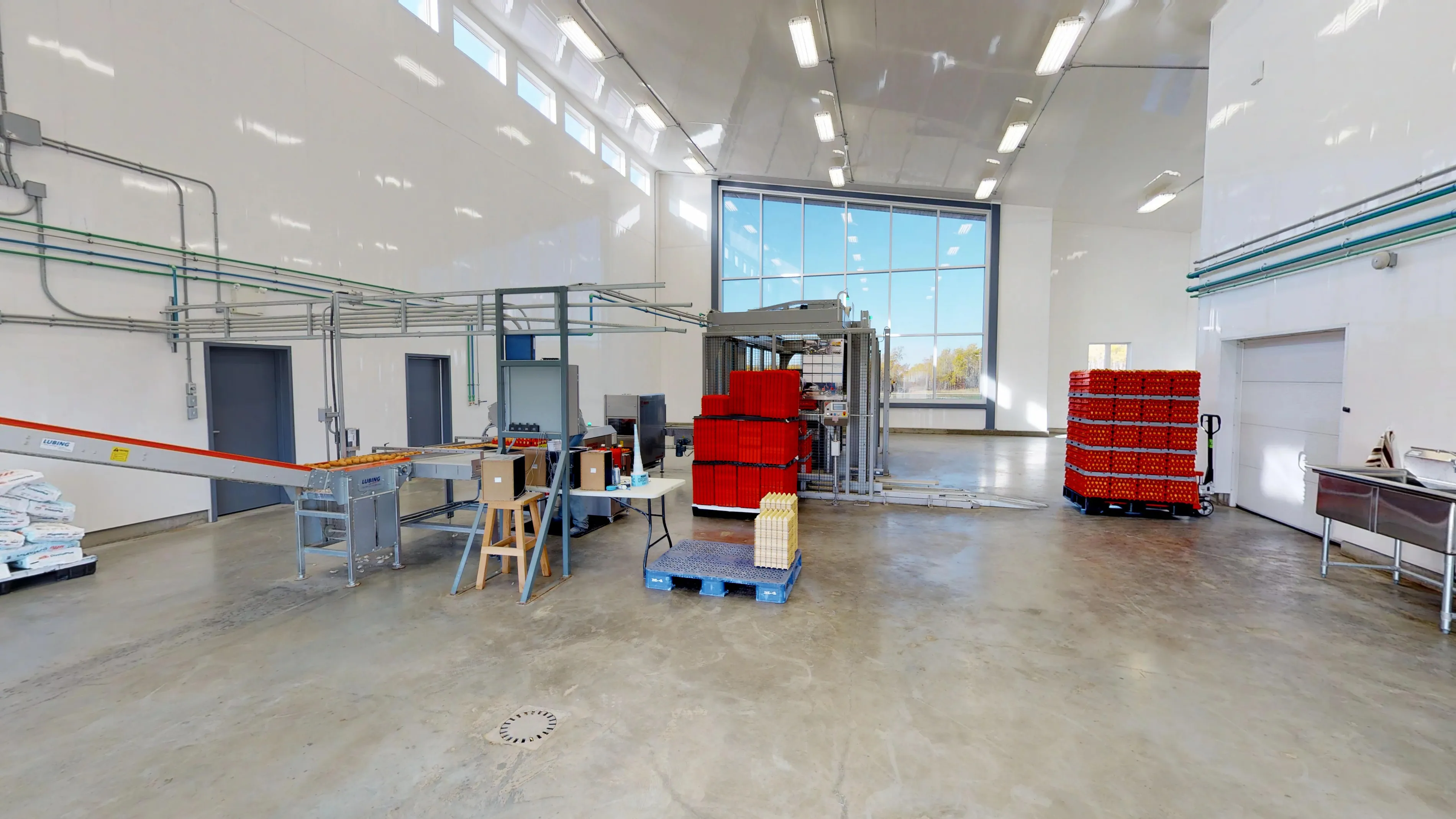 Bright, spacious industrial room with high ceilings and large windows. Stacks of red crates, a conveyor belt, equipment, and pallets are visible.