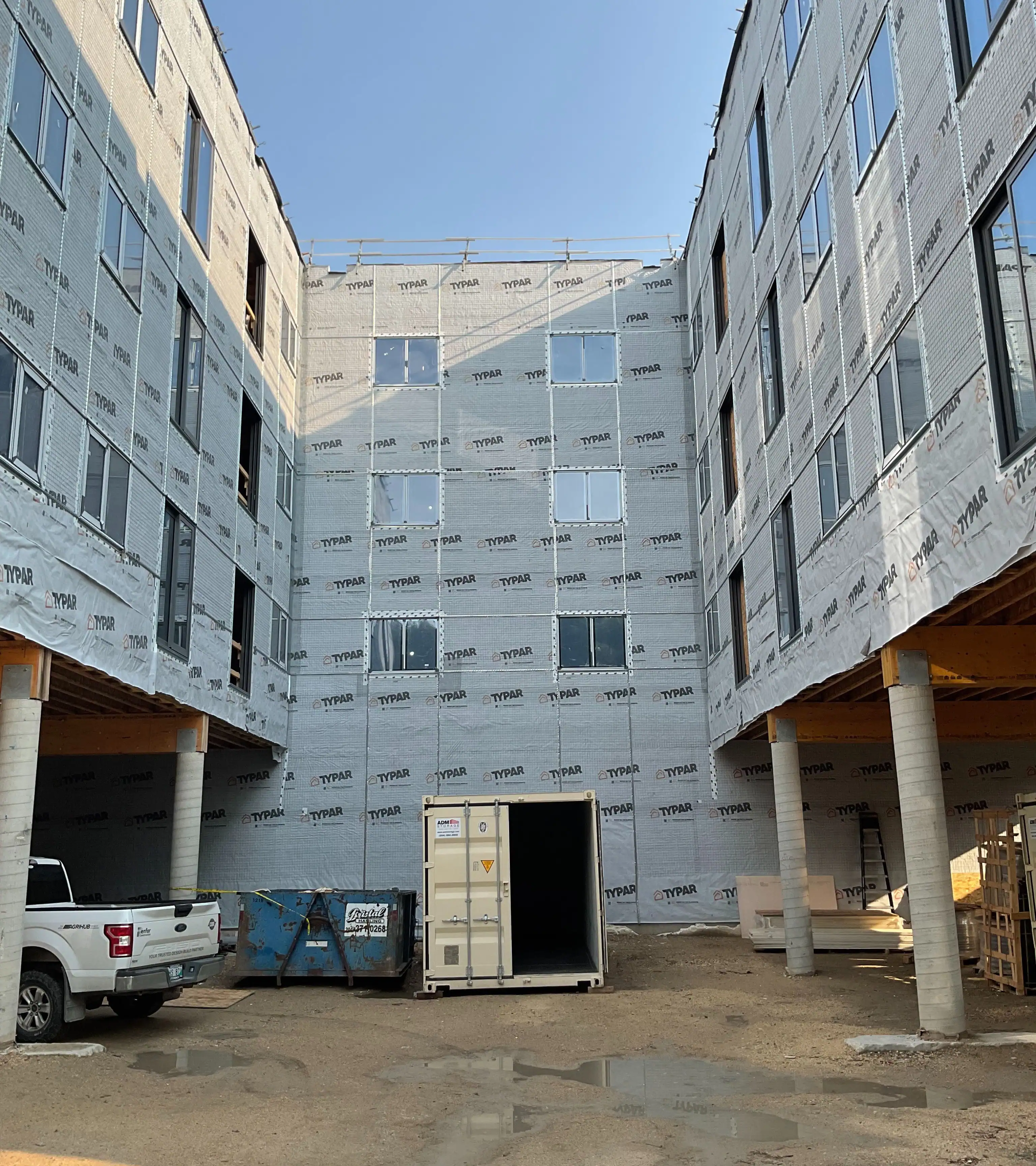 Construction site courtyard surrounded by four-story buildings wrapped in Typar material. The ground is dirt with a white truck and storage container visible.