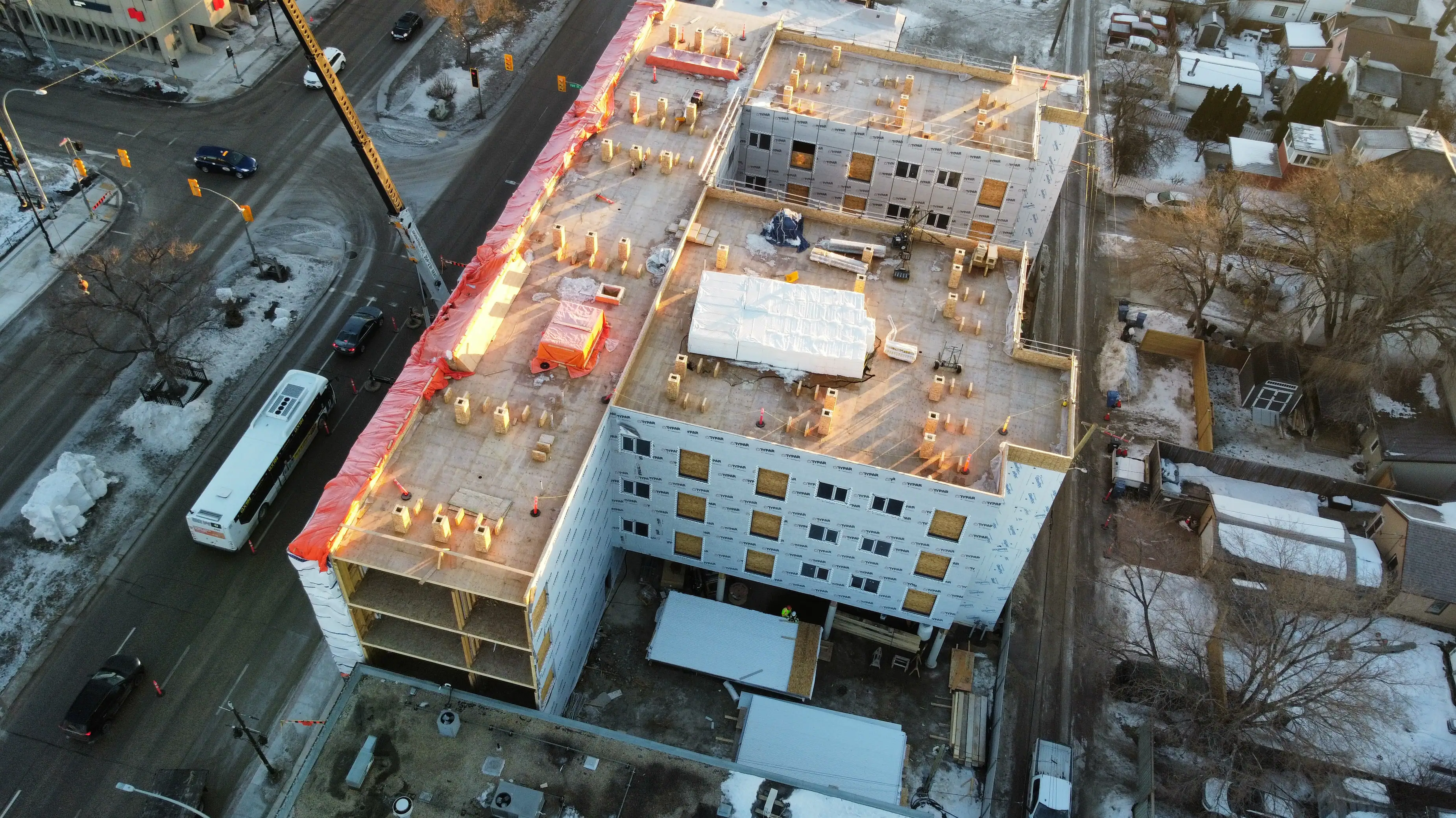 Aerial view of an unfinished multi-story building at a snowy intersection. Construction materials and a crane are visible under warm sunlight. Urban setting.