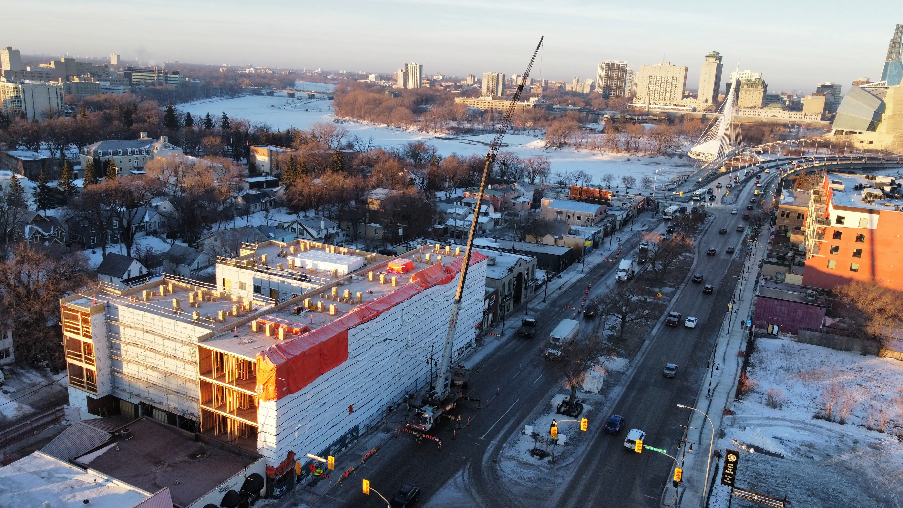 Aerial view of a snowy cityscape with a large building under construction in the foreground. A crane is present, and nearby roads are busy with cars.