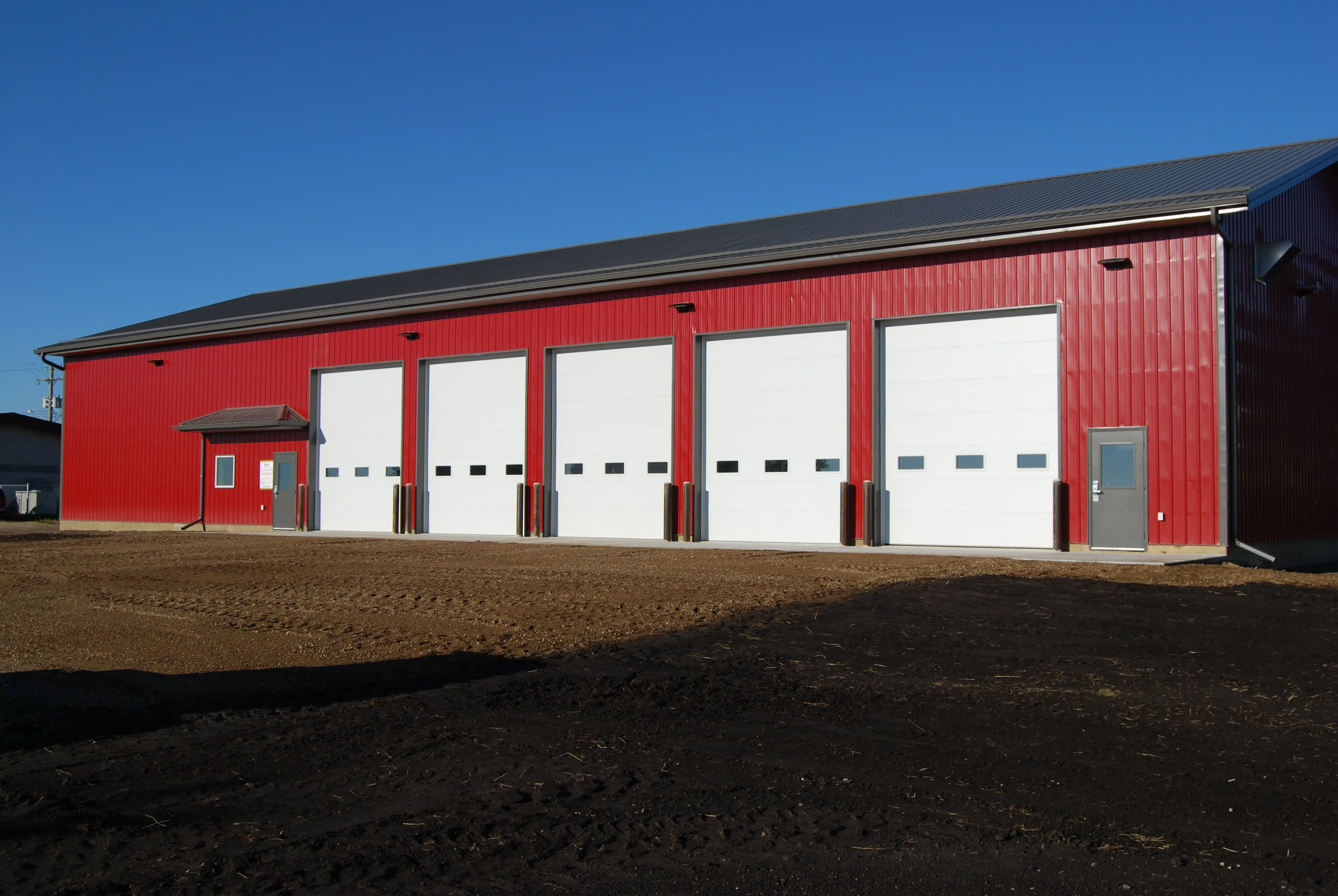 Red industrial building with five large white garage doors and two smaller entrance doors. The ground is dirt, under a clear blue sky.