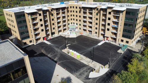 Aerial view of a newly constructed multi-storey apartment building with a U-shaped design, featuring light tan and dark exterior panels, multiple balconies, and rooftop mechanical units. The central courtyard and surrounding parking area are freshly paved, with construction still underway, equipment on-site, and landscaping not yet completed.