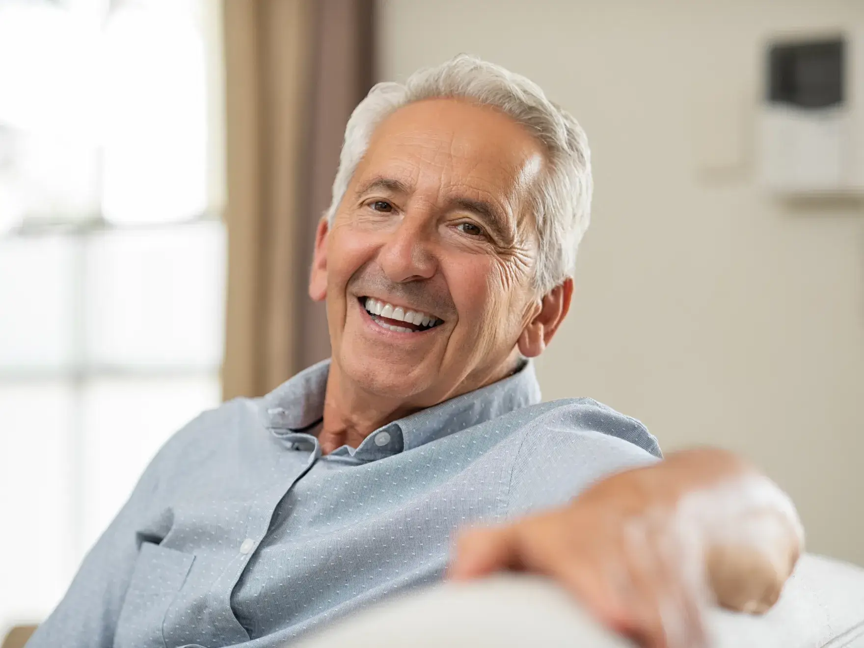 An older man with gray hair is sitting on a couch, smiling warmly at the camera.