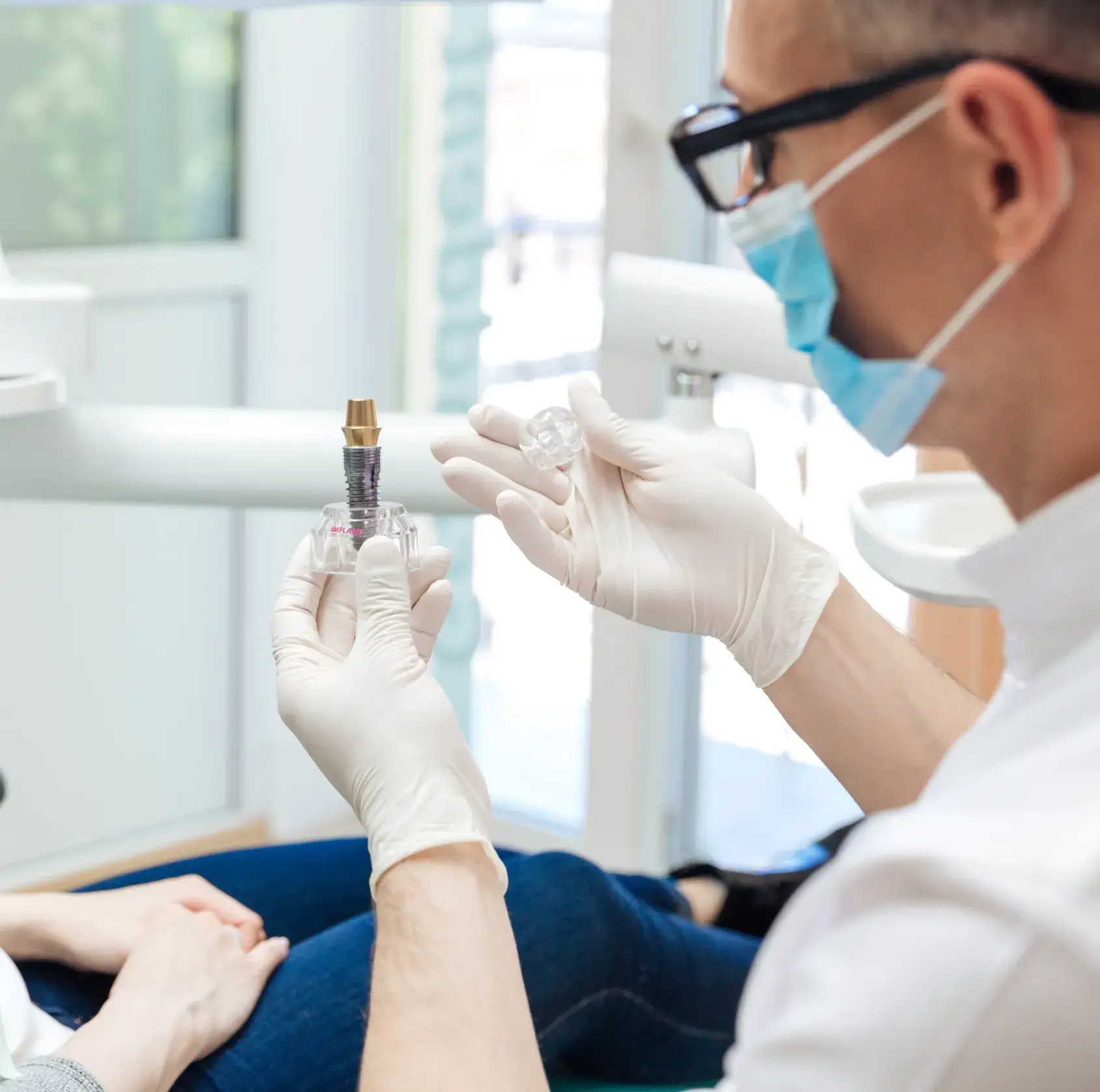 Dentist wearing gloves and mask shows a dental implant model to a seated patient.