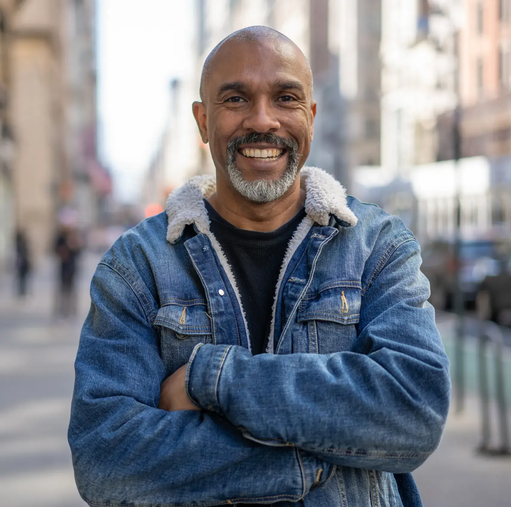 A man in a denim jacket stands on a city street, smiling with arms crossed.