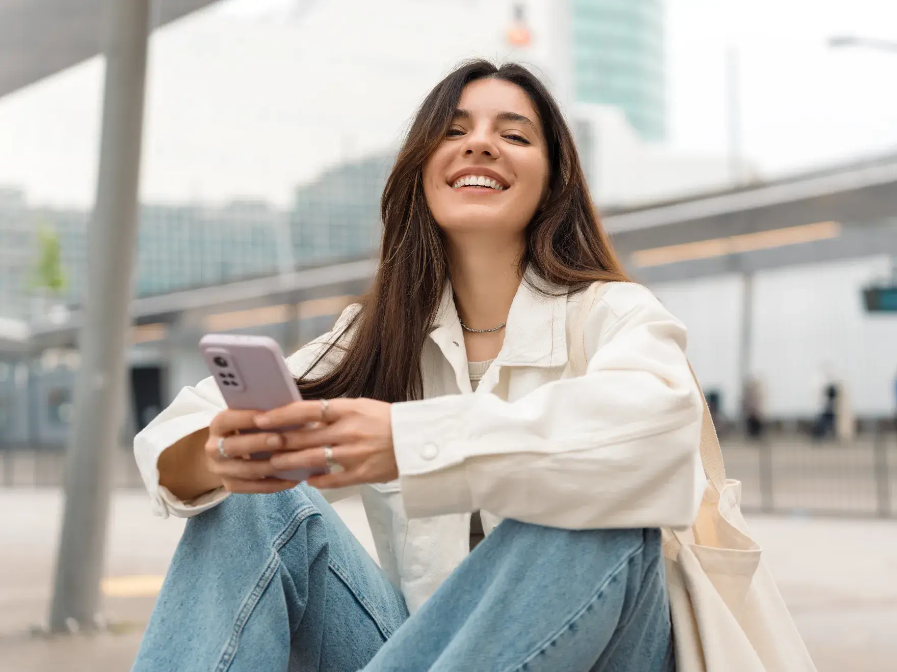 Woman sitting outdoors, smiling while holding a smartphone in both hands.