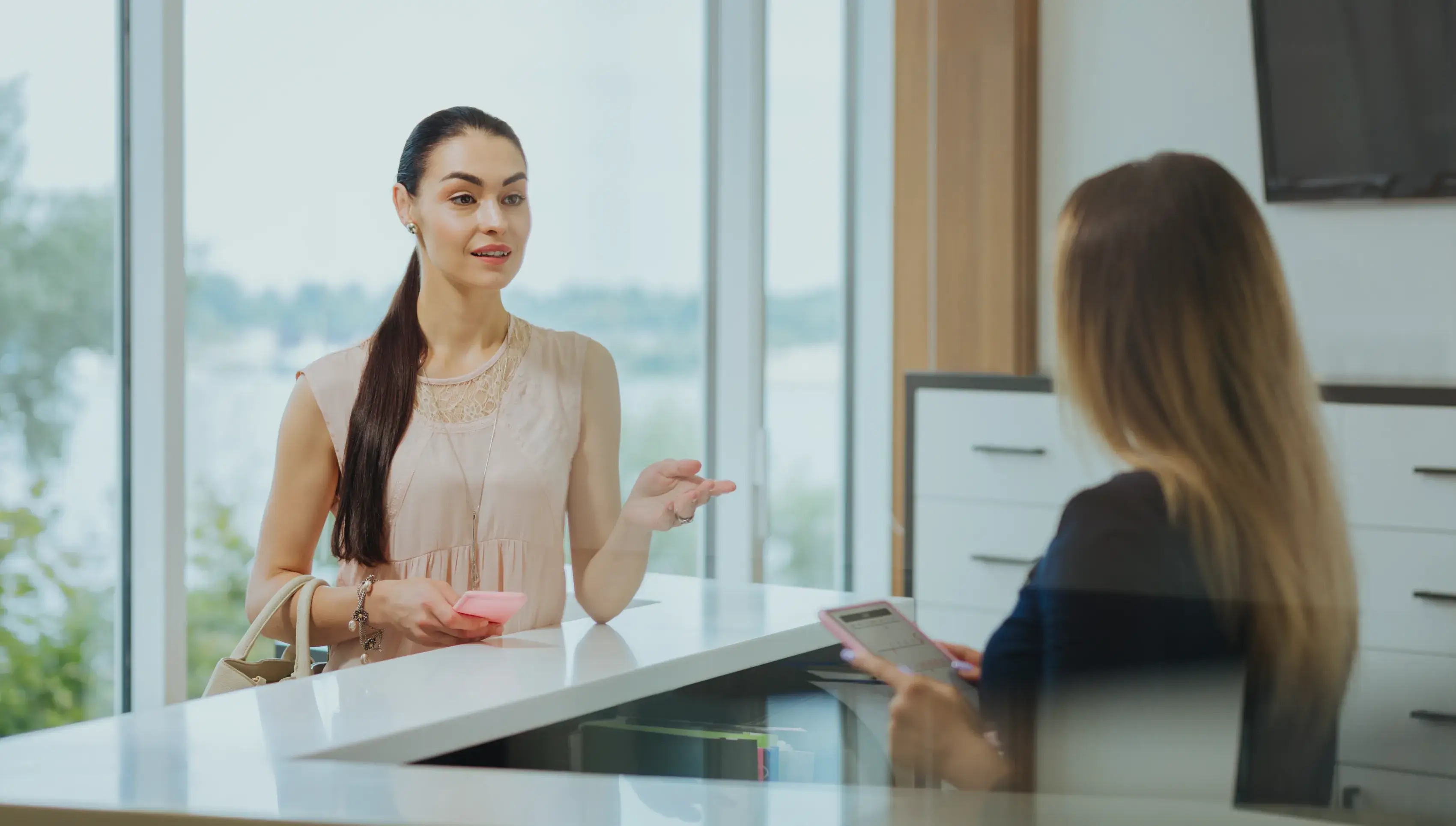 A woman stands at a reception desk, talking to a receptionist who is using a tablet.