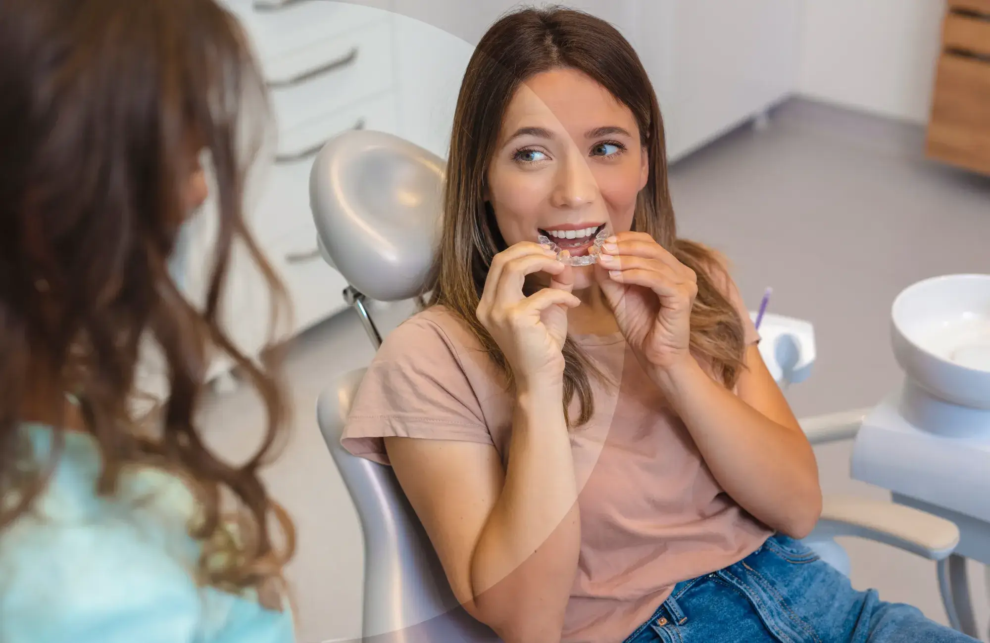 A woman in a dental chair holds and looks at a clear dental aligner, speaking to her dentist.