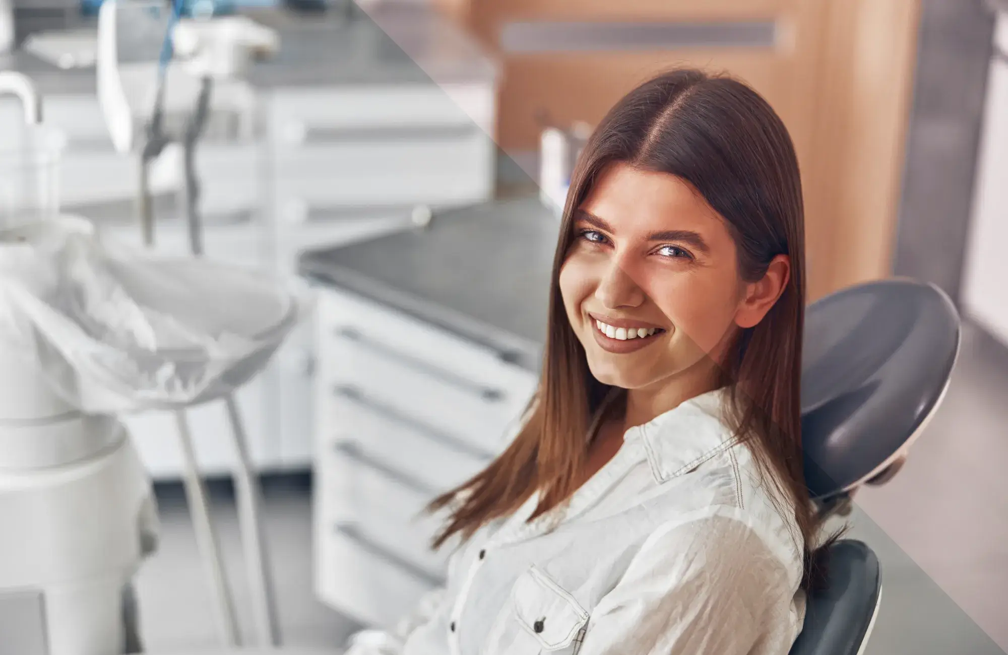 A woman with long brown hair sits smiling in a dentist's chair, wearing a white shirt.