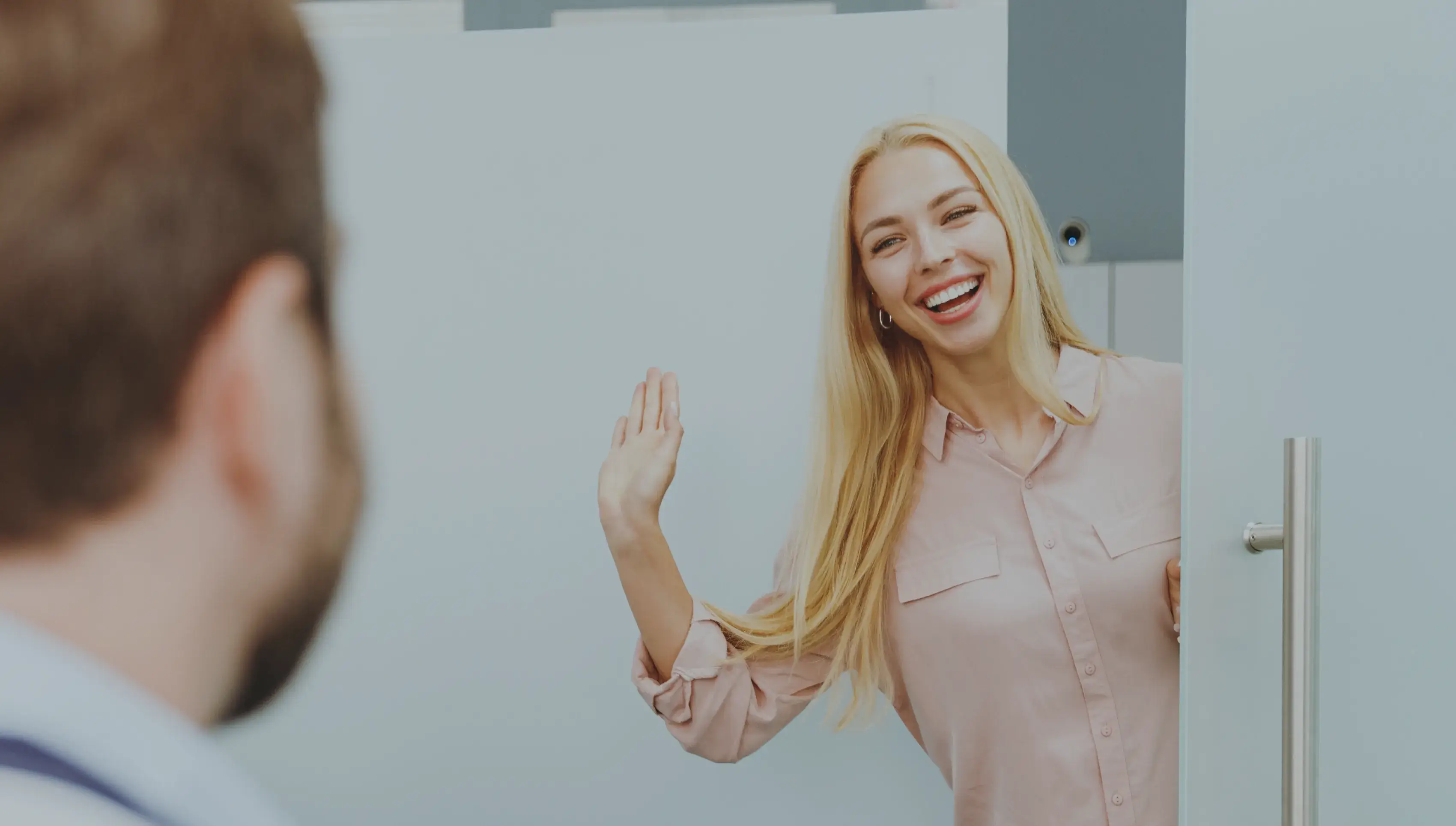 A woman with long blonde hair smiles and waves as she enters a room.