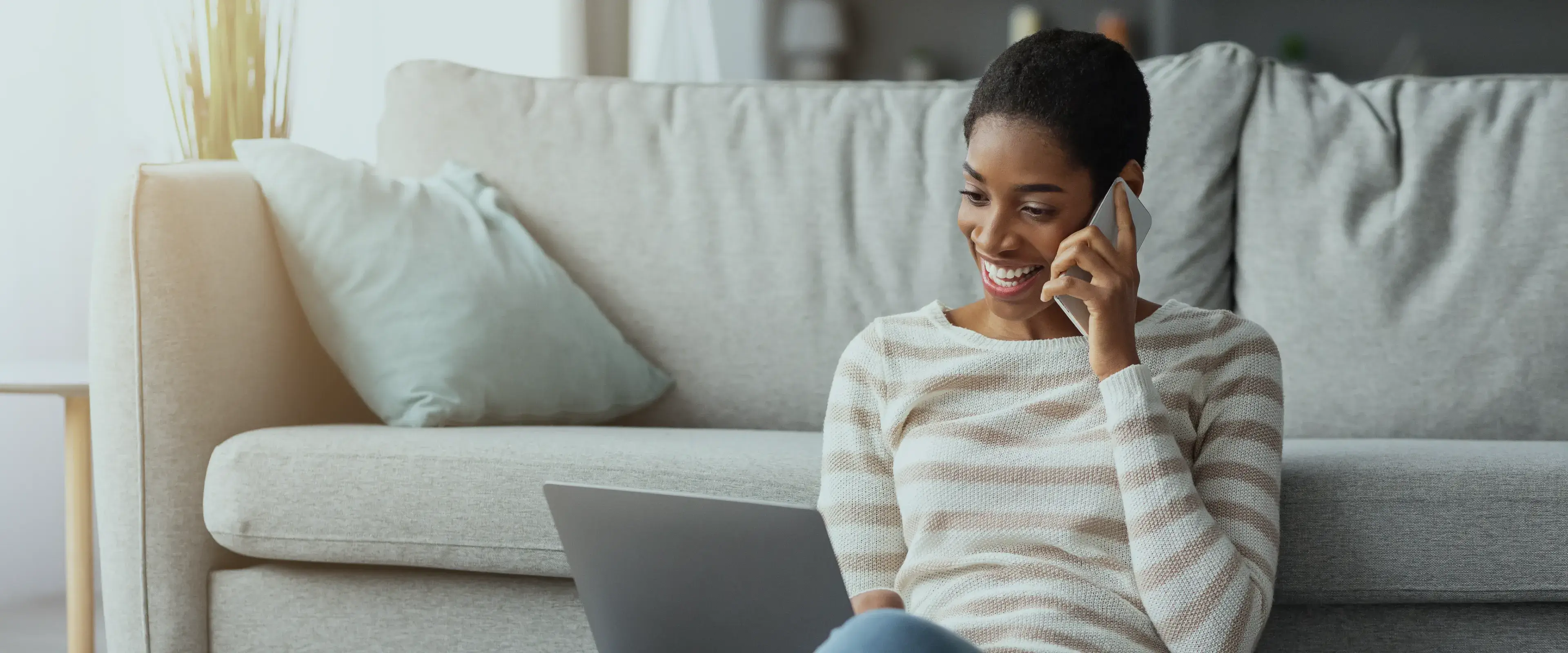 A woman sits on a couch, smiling while talking on her phone with a laptop nearby.