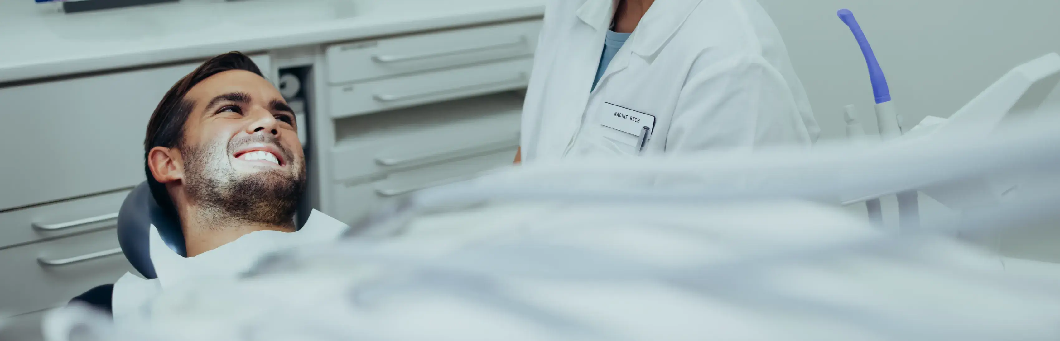 A person in a dental chair smiles while a dentist prepares for a procedure.
