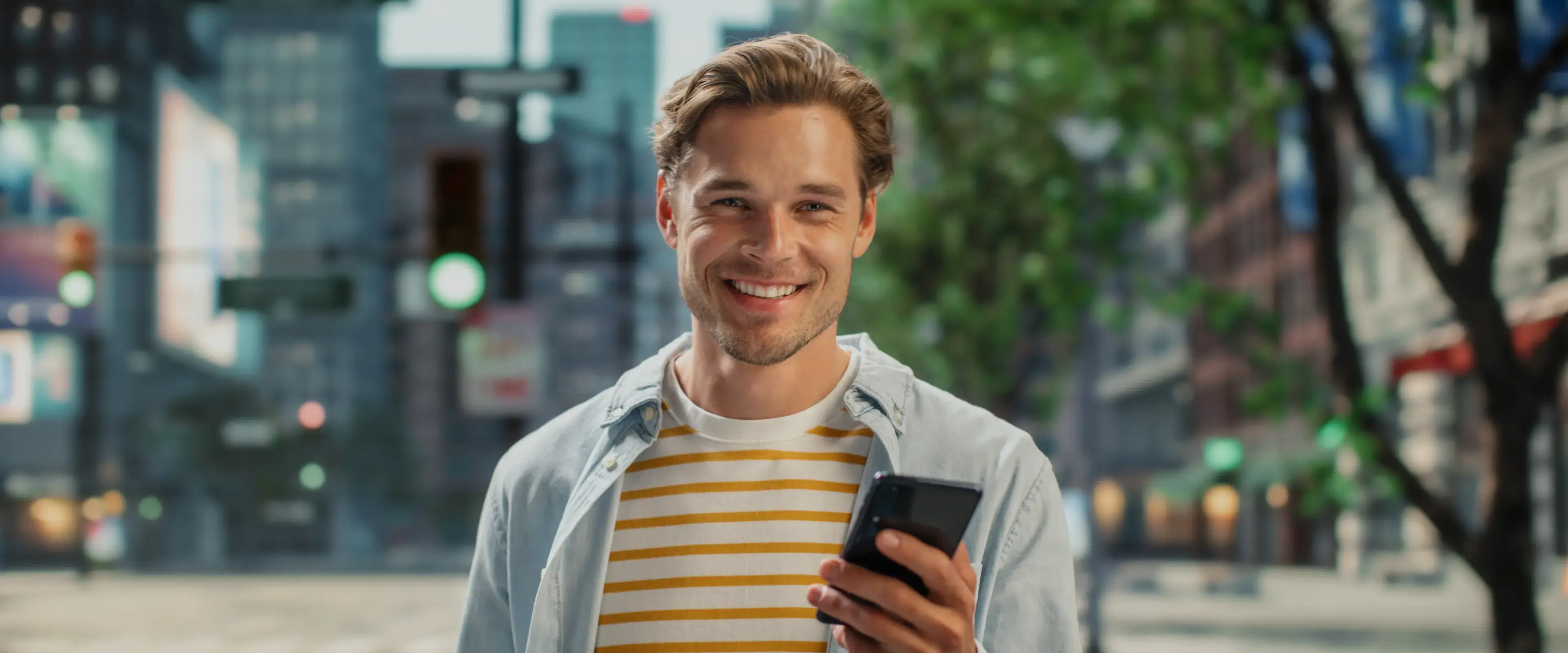 A man is smiling while holding a smartphone on a city street.