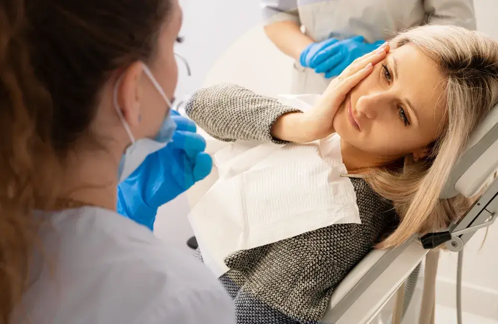 A woman in a dentist's chair holds her cheek, looking at a dentist.