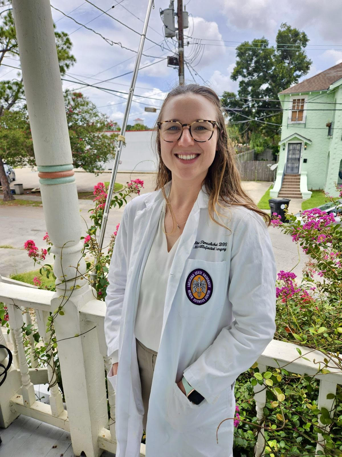 Mari-Alina Timoshchuk in a white coat on a porch in New Orleans with purple flowers in the background