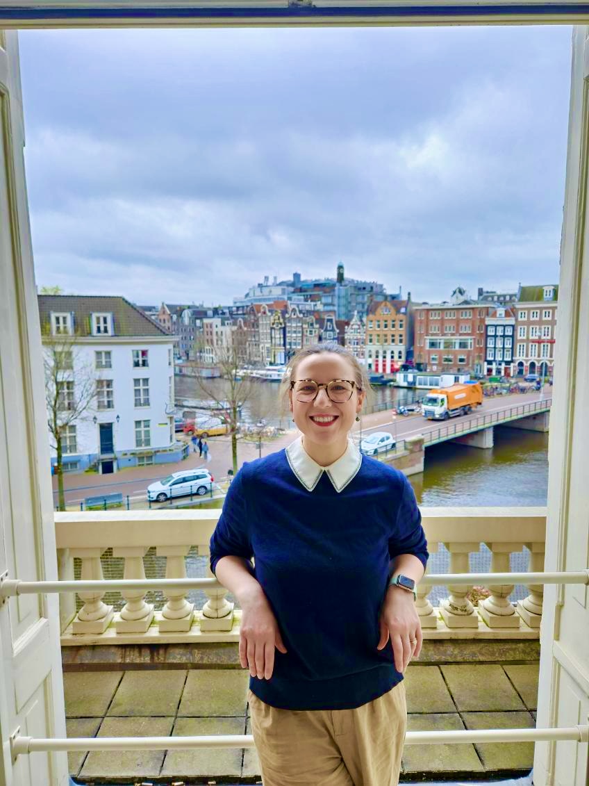 Mari-Alina Timoshchuk in Amsterdam smiling in a dark blue sweater with the Amsterdam canal and houses in the background. It's a sunny day. 