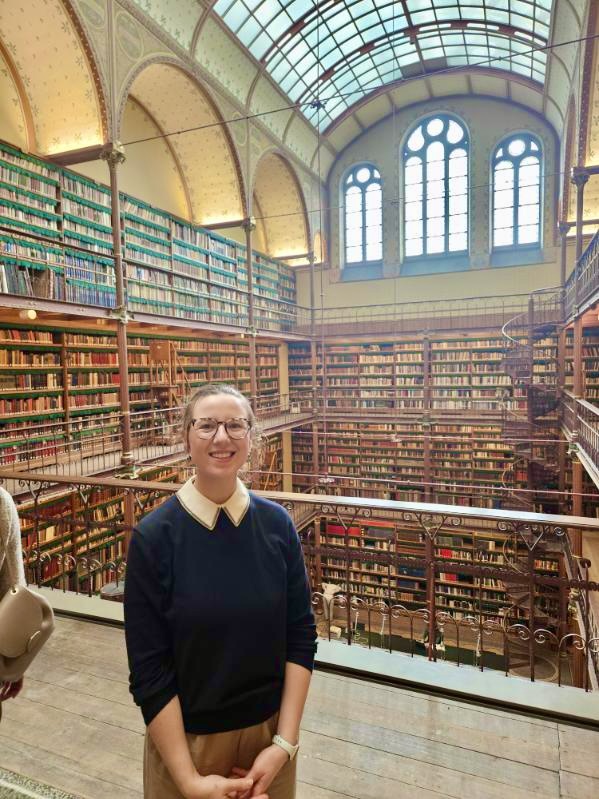 Mari-Alina Timoshchuk at a library in Prague wearing a blue sweater and smiling. There is a wall of books behind her and beautiful large windows. 