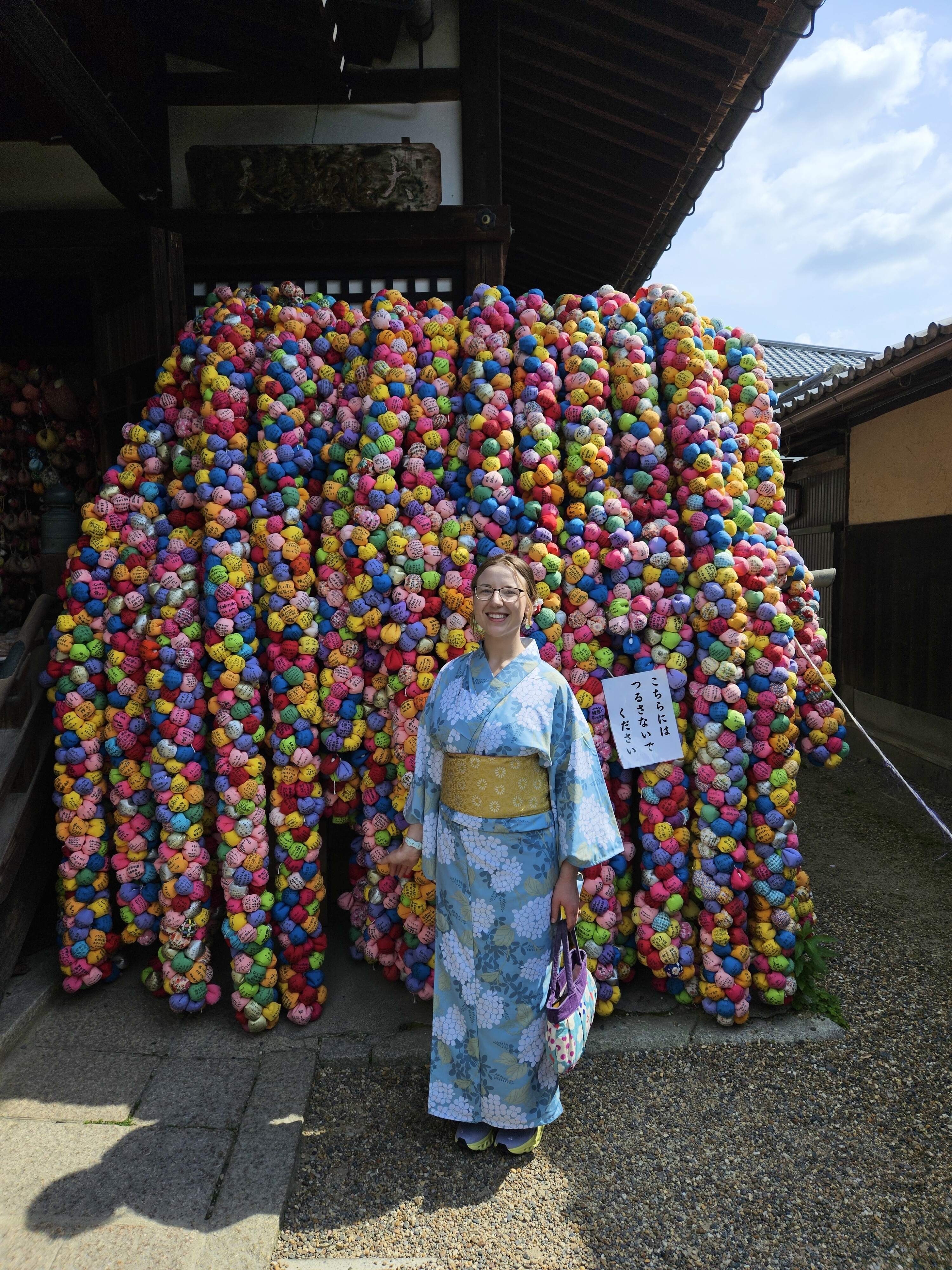 Mari-Alina Timoshchuk in front of the monkey temple in Kyoto Japan in a blue kimono smiling. A colorful wall of kids toys is behind her. 