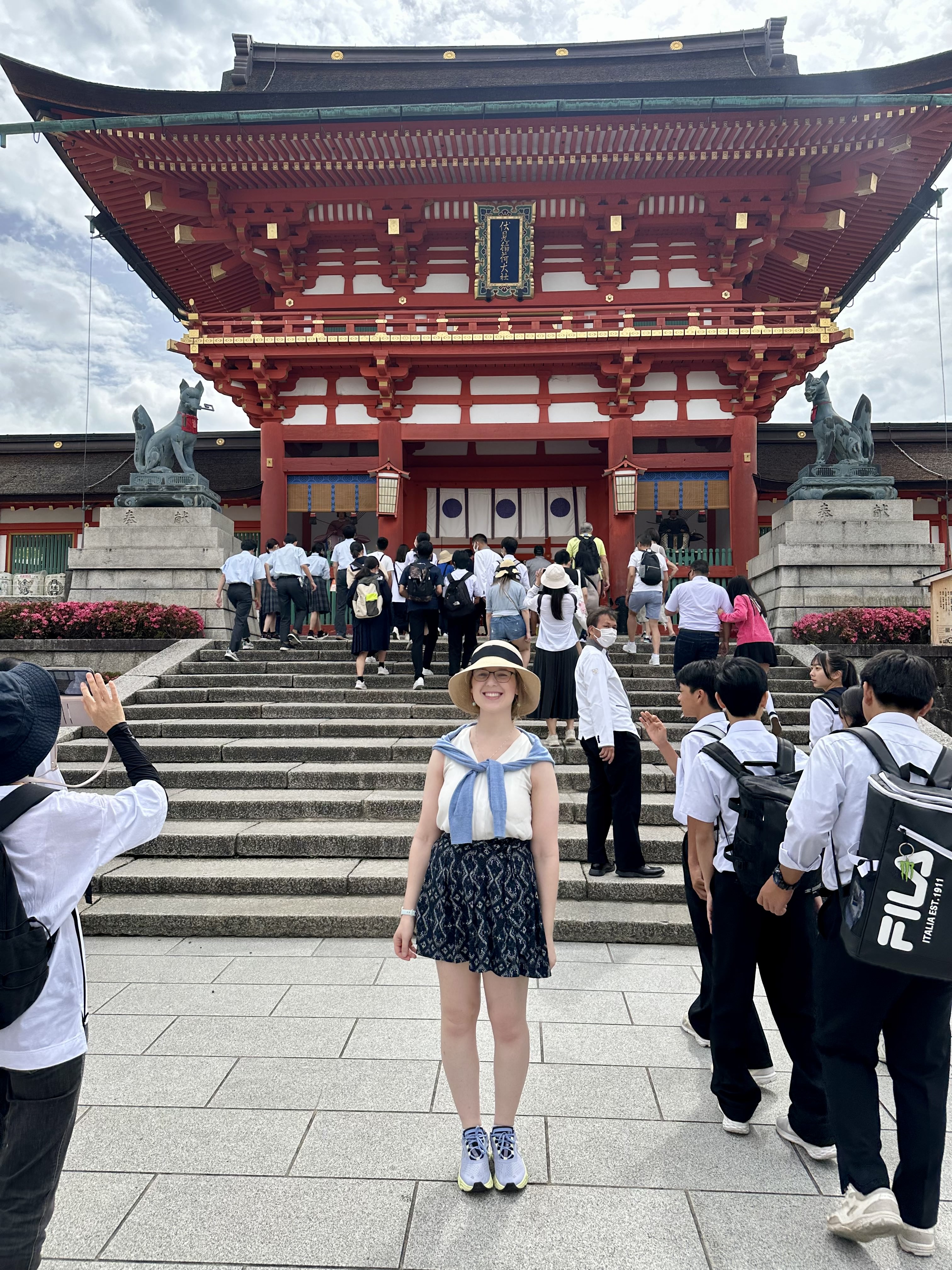 Mari-Alina Timoshchuk in front of Fushimi Inari wearing a straw hat, skirt, and blouse. School children are in the background and it's a sunny day. She is smiling. 