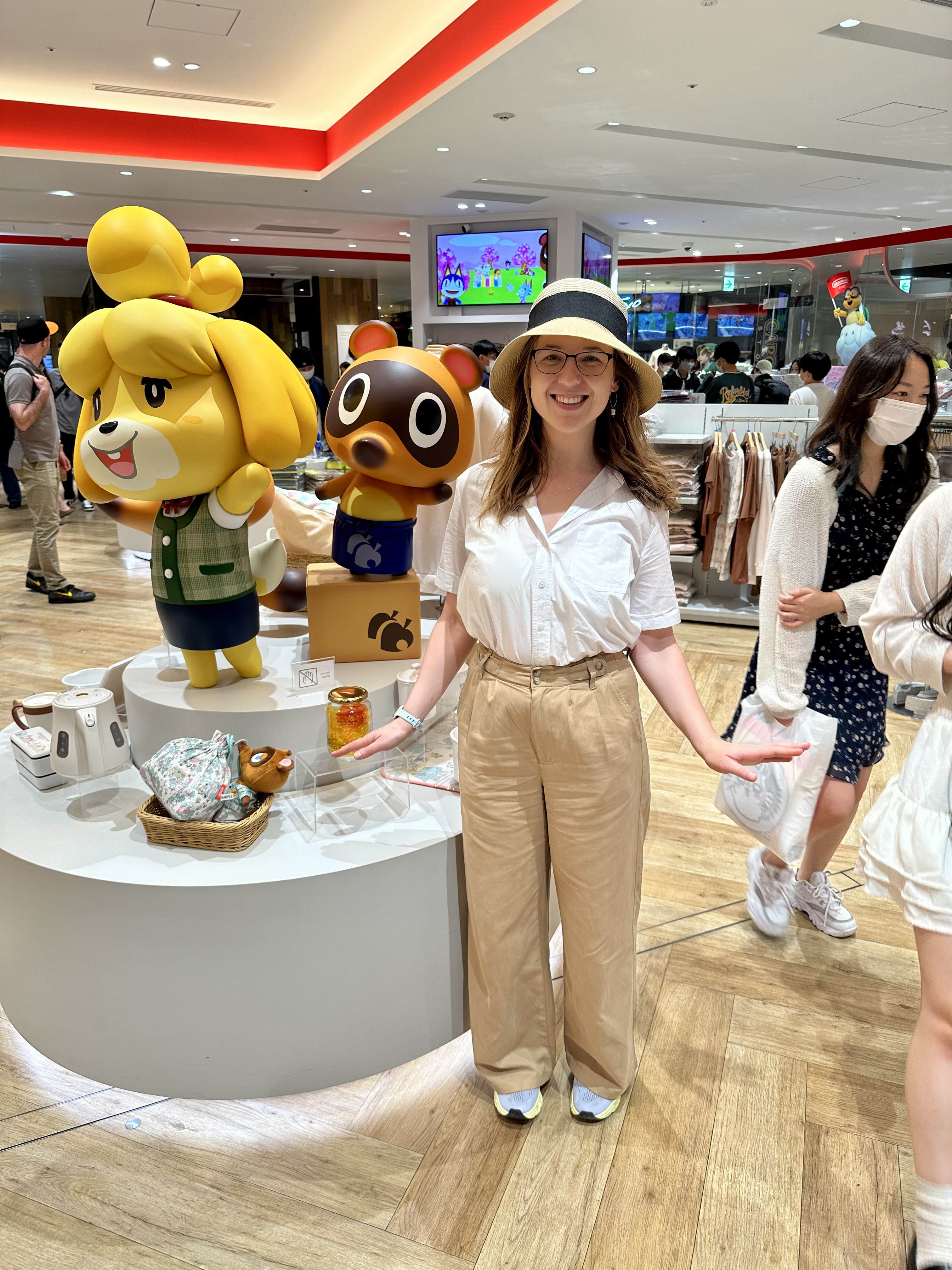 Mari-Alina Timoshchuk in the Nintendo store in Japan next to a life sized Animal Crossing Character. She is smiling. She is wearing a hat, brown pants, and a white blouse.