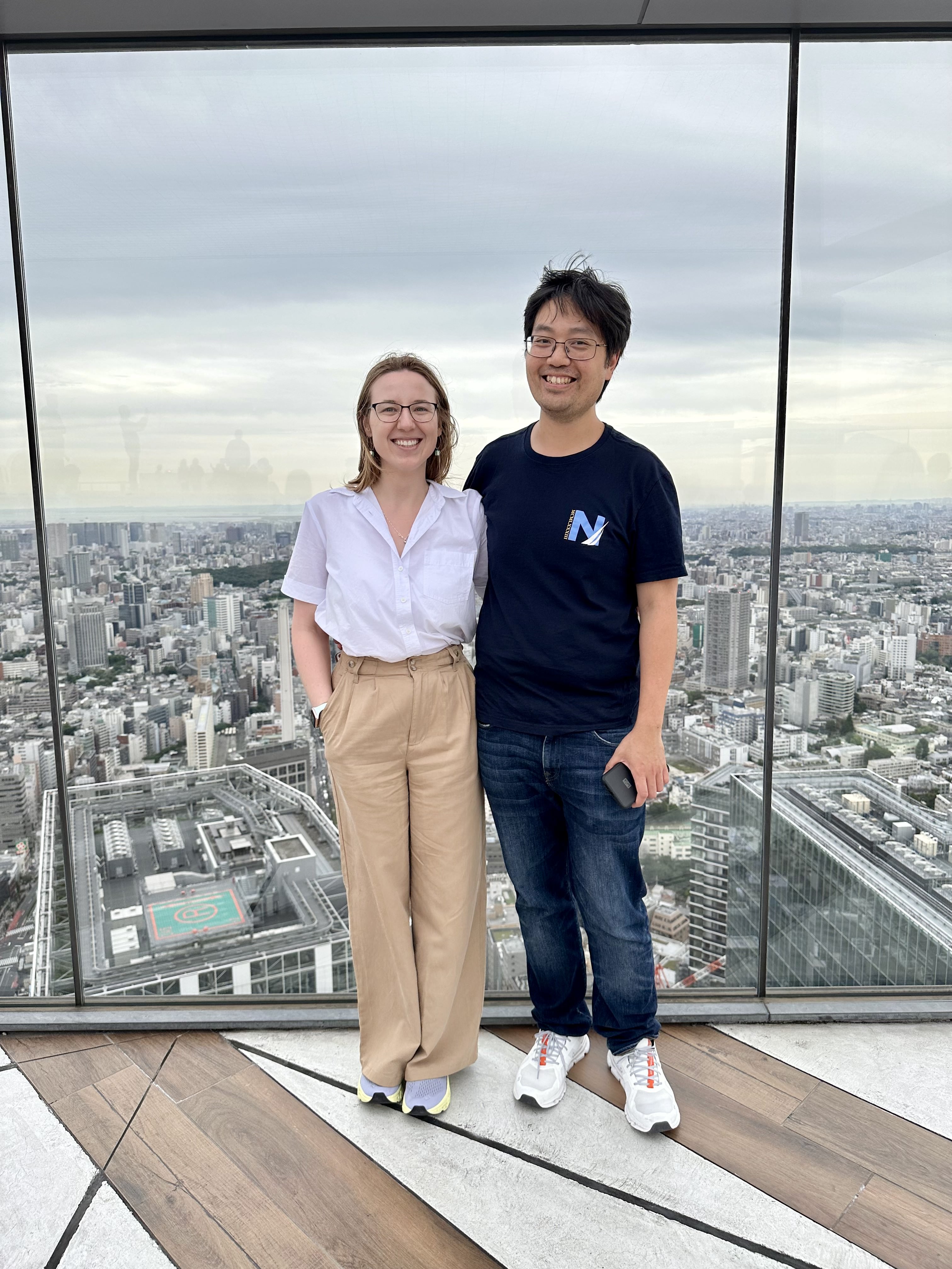 Mari-Alina Timoshchuk and Cassidy Sung on top of the highest tower in Tokyo with the wind blowing. They are smiling. It's a beautiful day. There is Tokyo behind them.