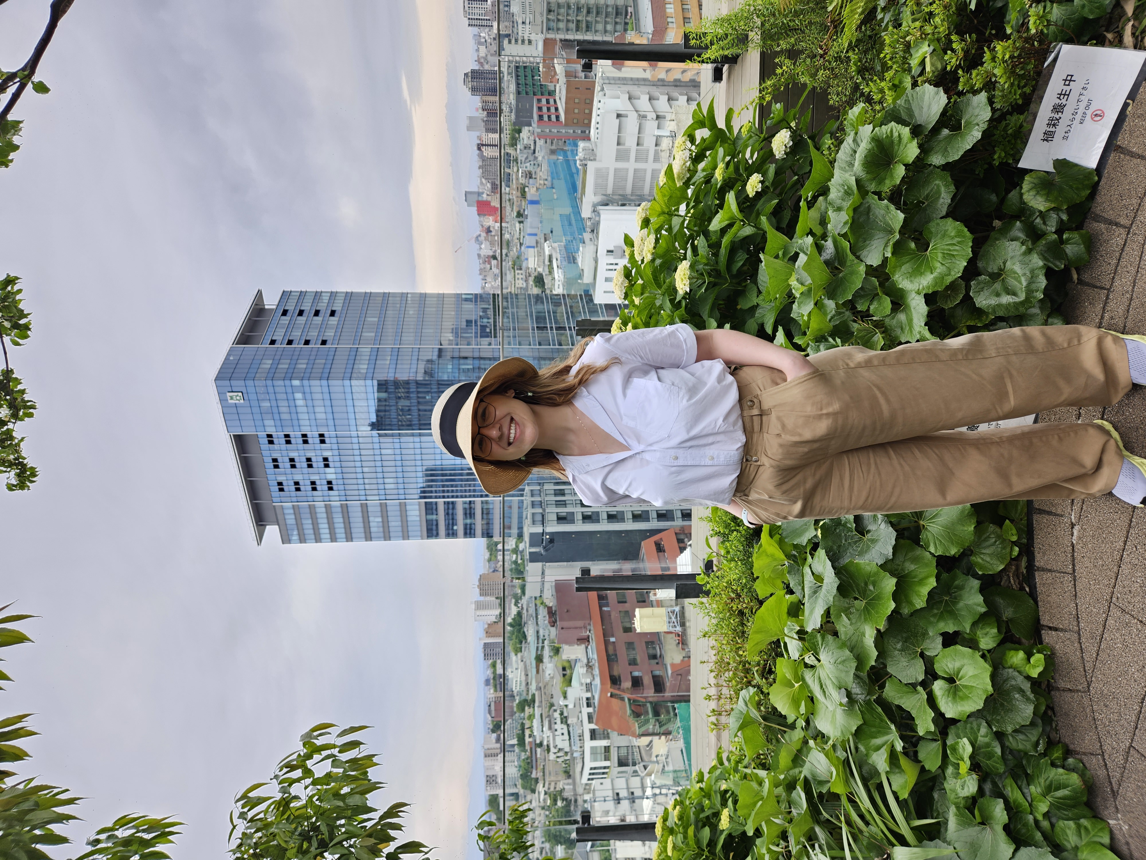 Mari-Alina Timoshchuk at a skyscraper in Tokyo with Tokyo in the background. She is wearing a brown hat and brown pants with a white blouse and smiling.