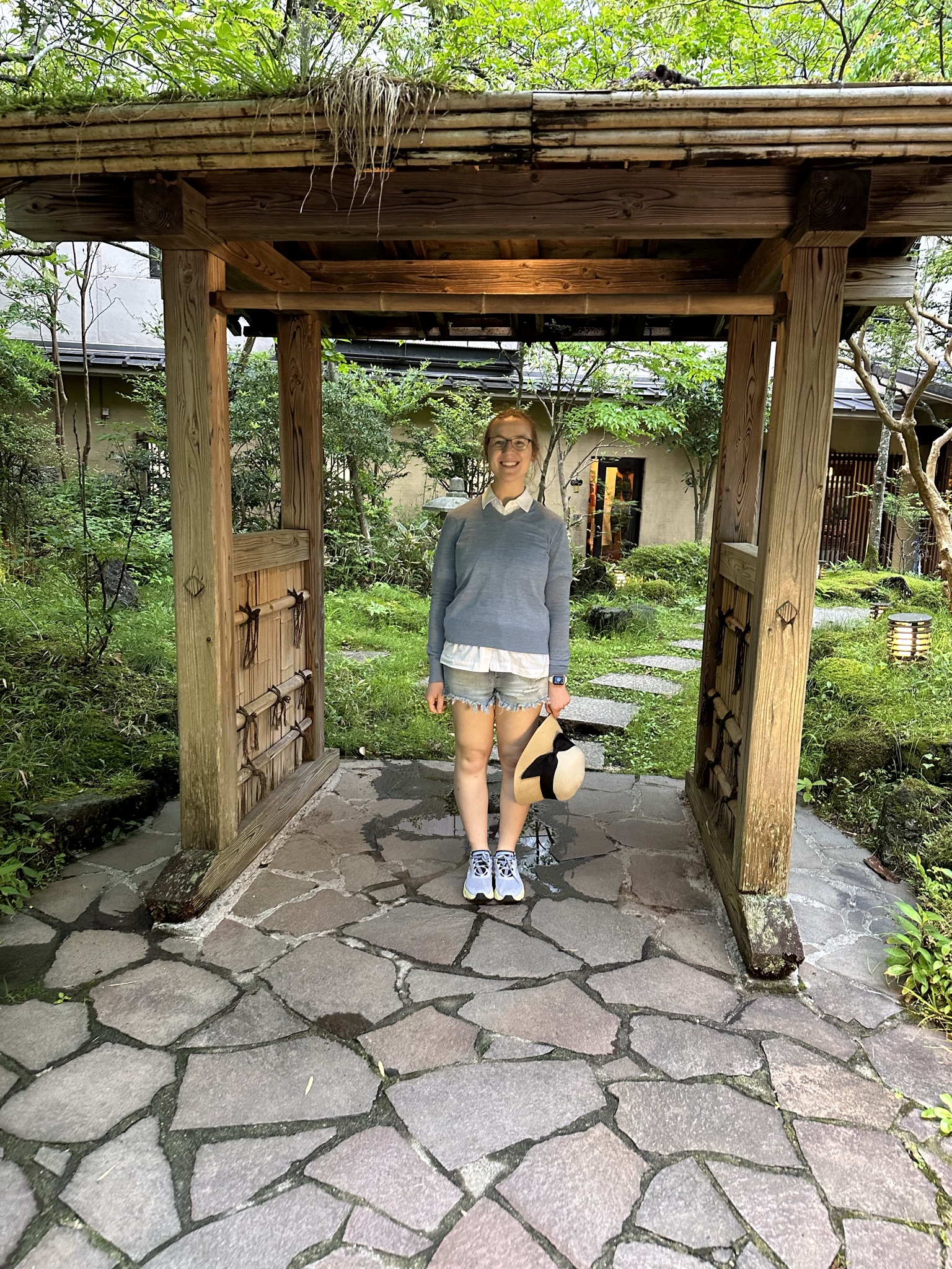 Mari-Alina Timoshchuk in front of a Japanese inn with a blue sweater and holding a hat. She is smiling. She is under the entrance which is wooden. There is a forest and a walkway in the background. 
