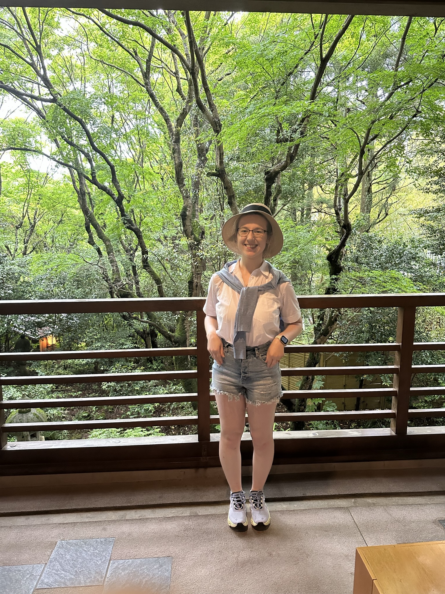 Mari-Alina Timoshchuk on a Japanese balcony with a forest in the background. She is wearing a hat and a blue sweater with shorts. 