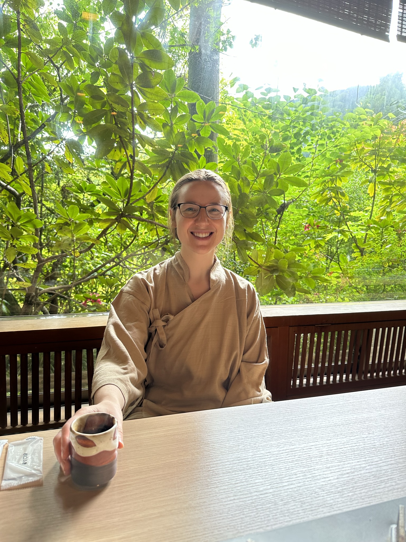 Mari-Alina Timoshchuk sitting at a table drinking tea and smiling. There is a Japanese forest in the background. 