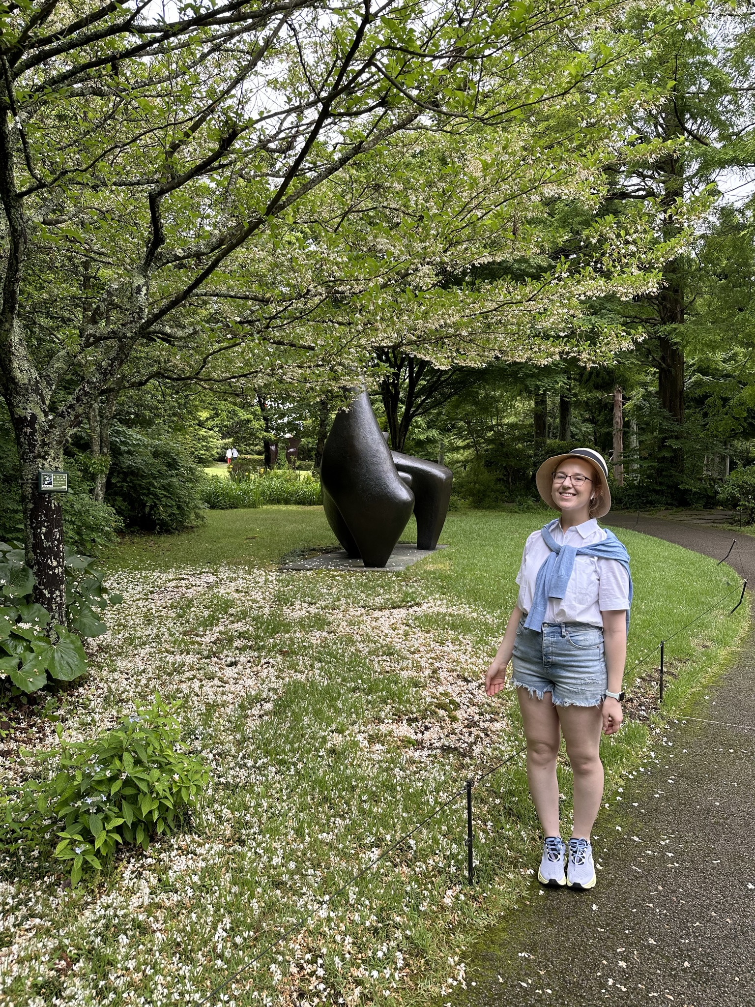 Mari-Alina Timoshchuk in the statue garden in Hakone next to a cherry blossom tree and a sculpture. She is wearing a hat, white blouse, and shorts. She is smiling. 