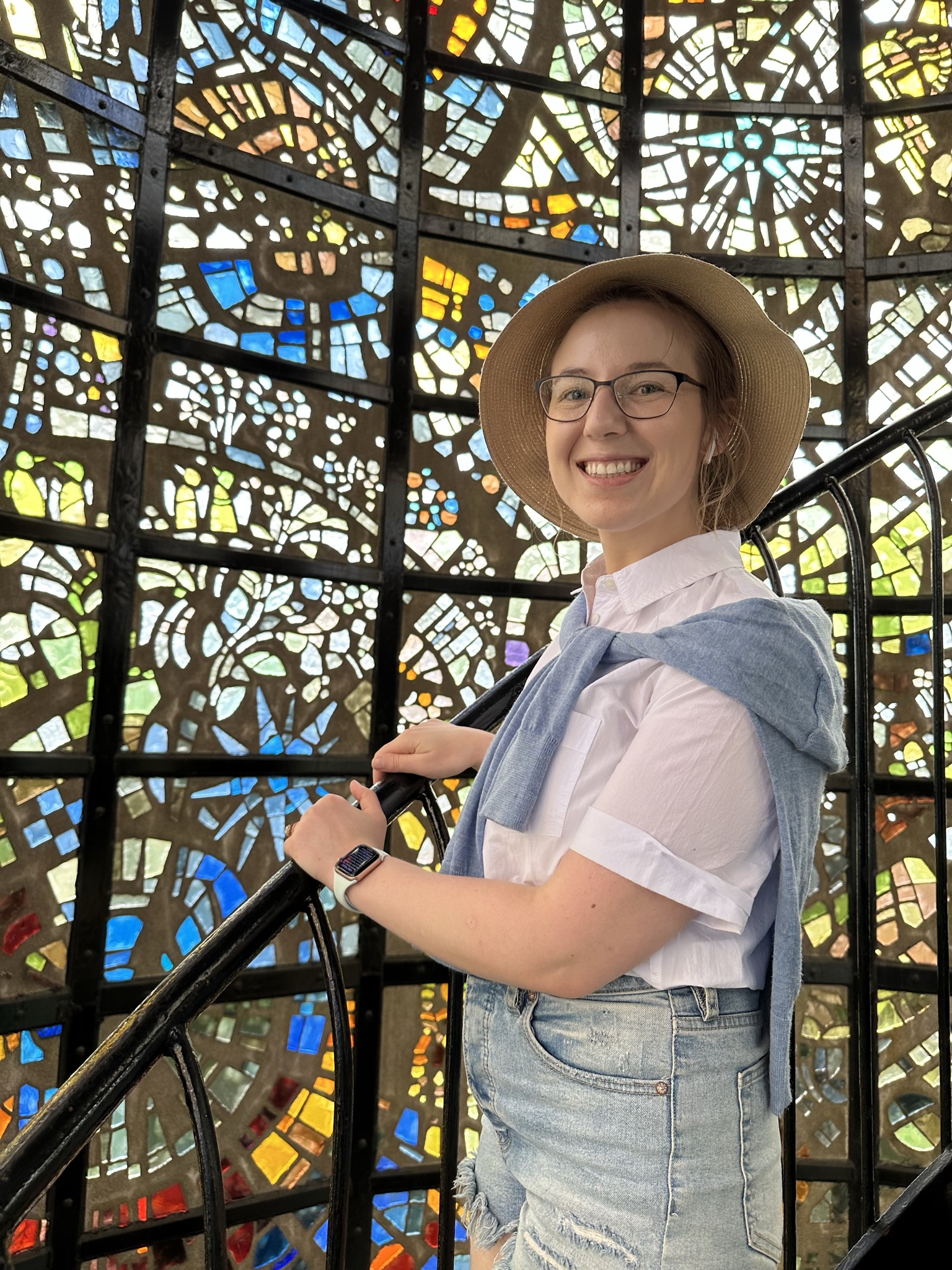 Mari-Alina Timoshchuk at the Symphony Structure in Hakone Japan with a wall of stained glass behind her. She is smiling and wearing a wide brimmed hat. 