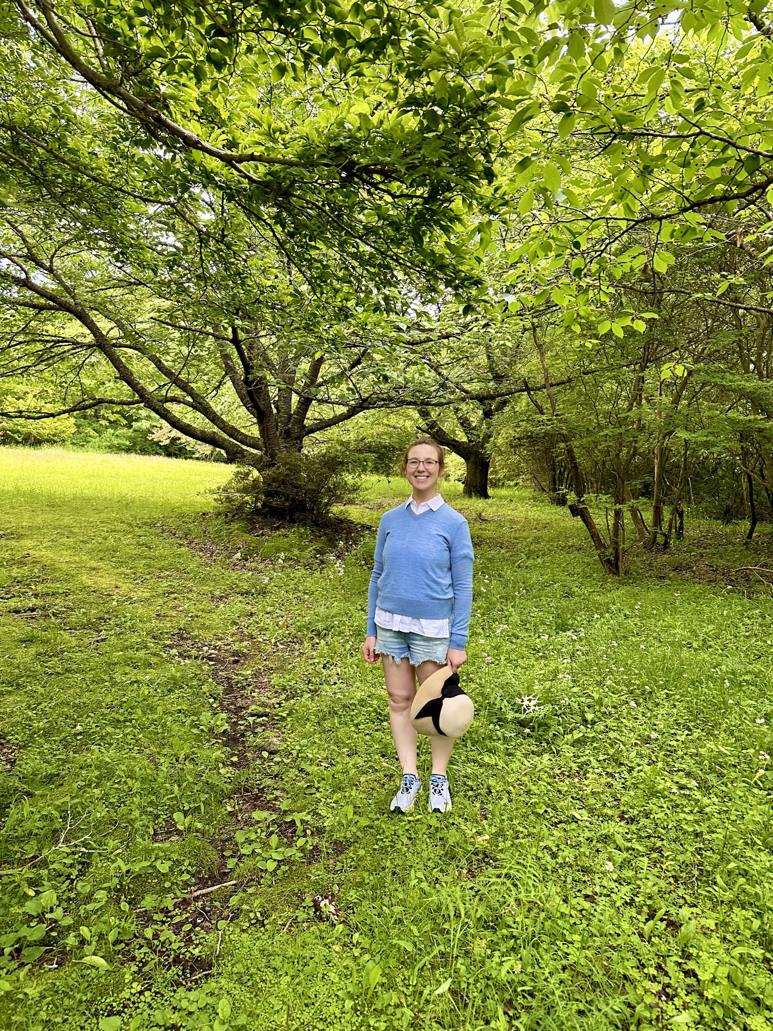 Mari-Alina Timoshchuk in a Japanese forest holding a wide brimmed hat in a blue sweater