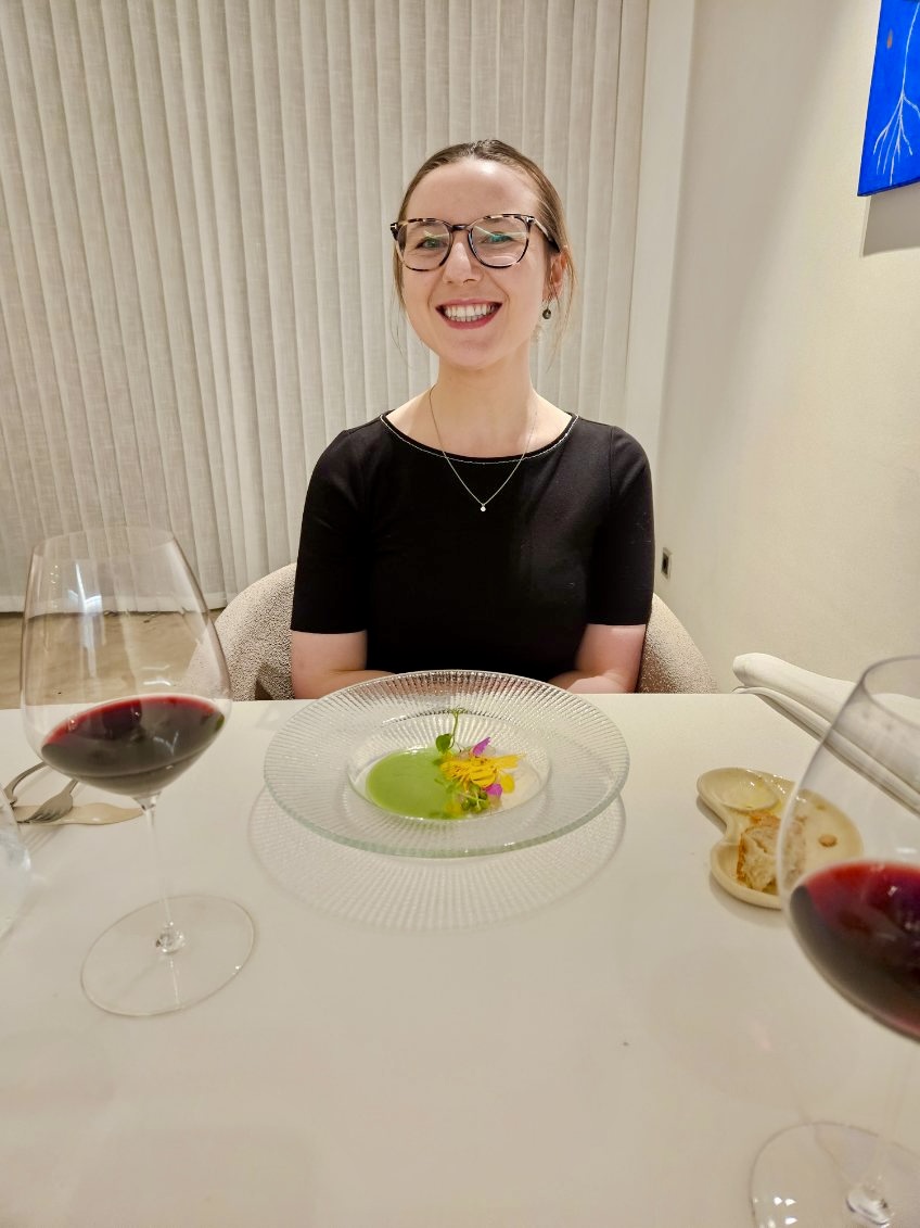 Mari-Alina Timoshchuk sitting at a dinner table in Madrid Spain next to a glass of red wine. She is smiling and wearing glasses. She is wearing a black dress. 