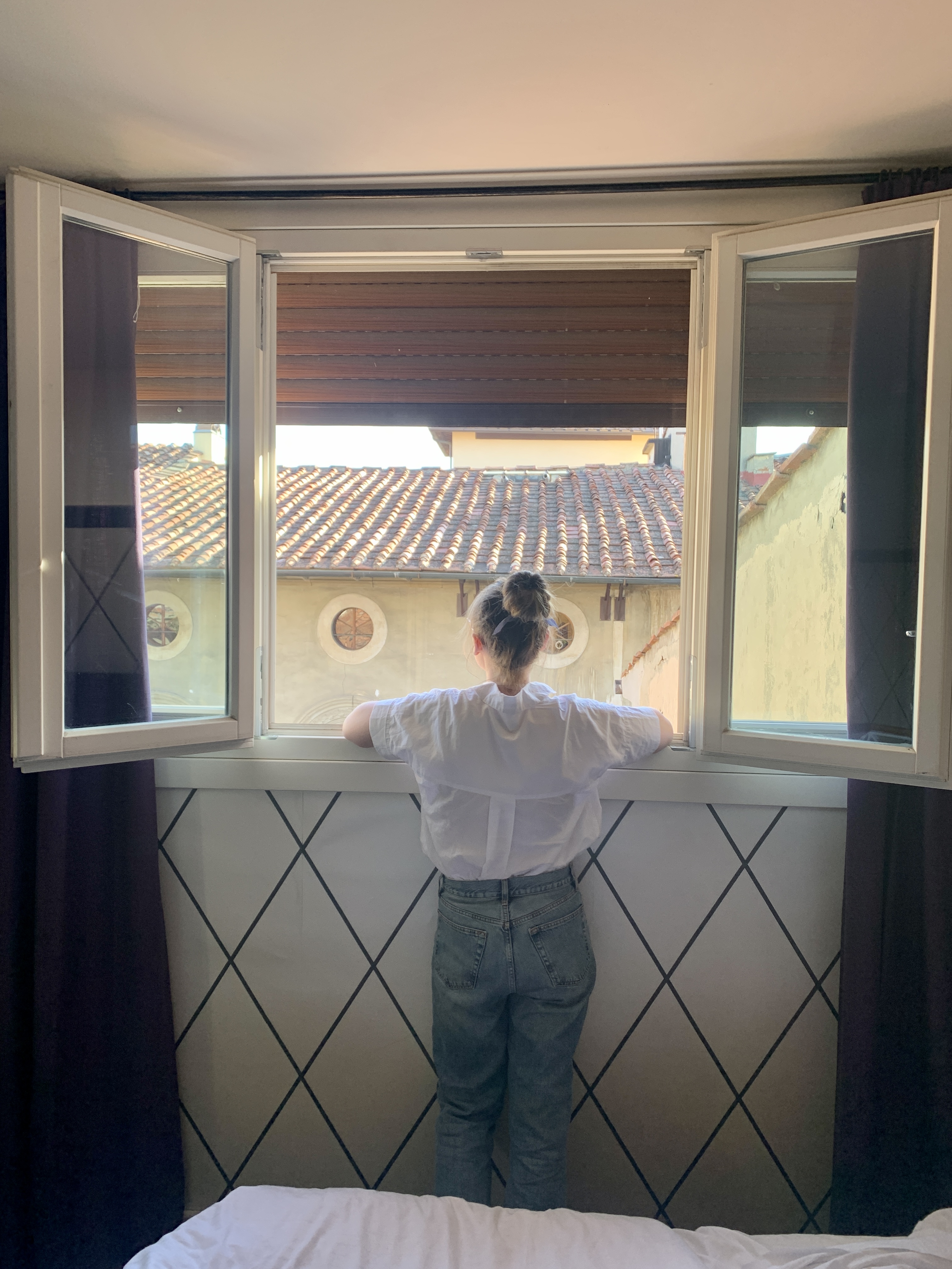 Mari-Alina Timoshchuk looking out a window in a room in Florence Italy. She is wearing a white blouse and jeans. Her hair is in a bun. 